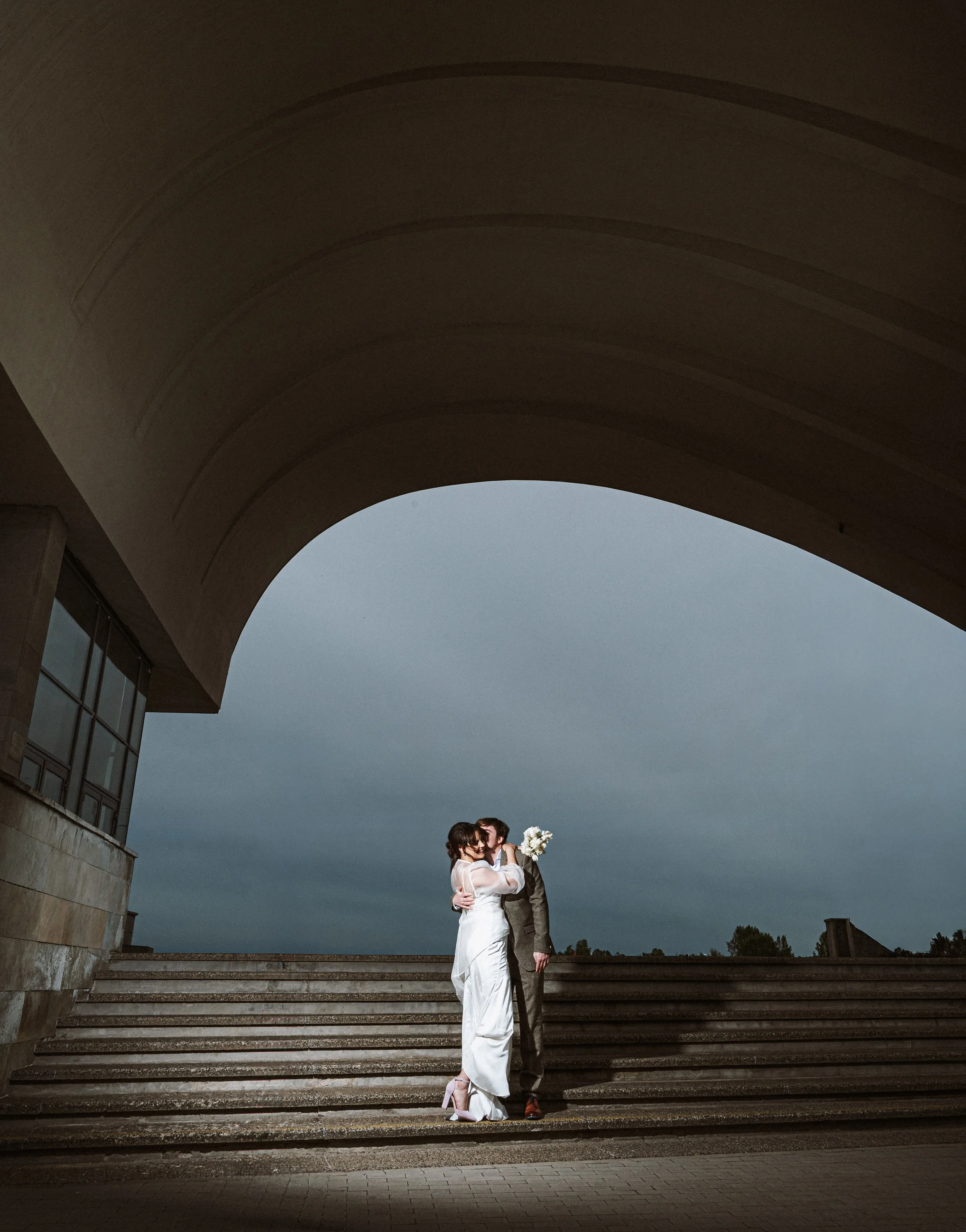 Couple embracing on steps outdoors under a large concrete arch.