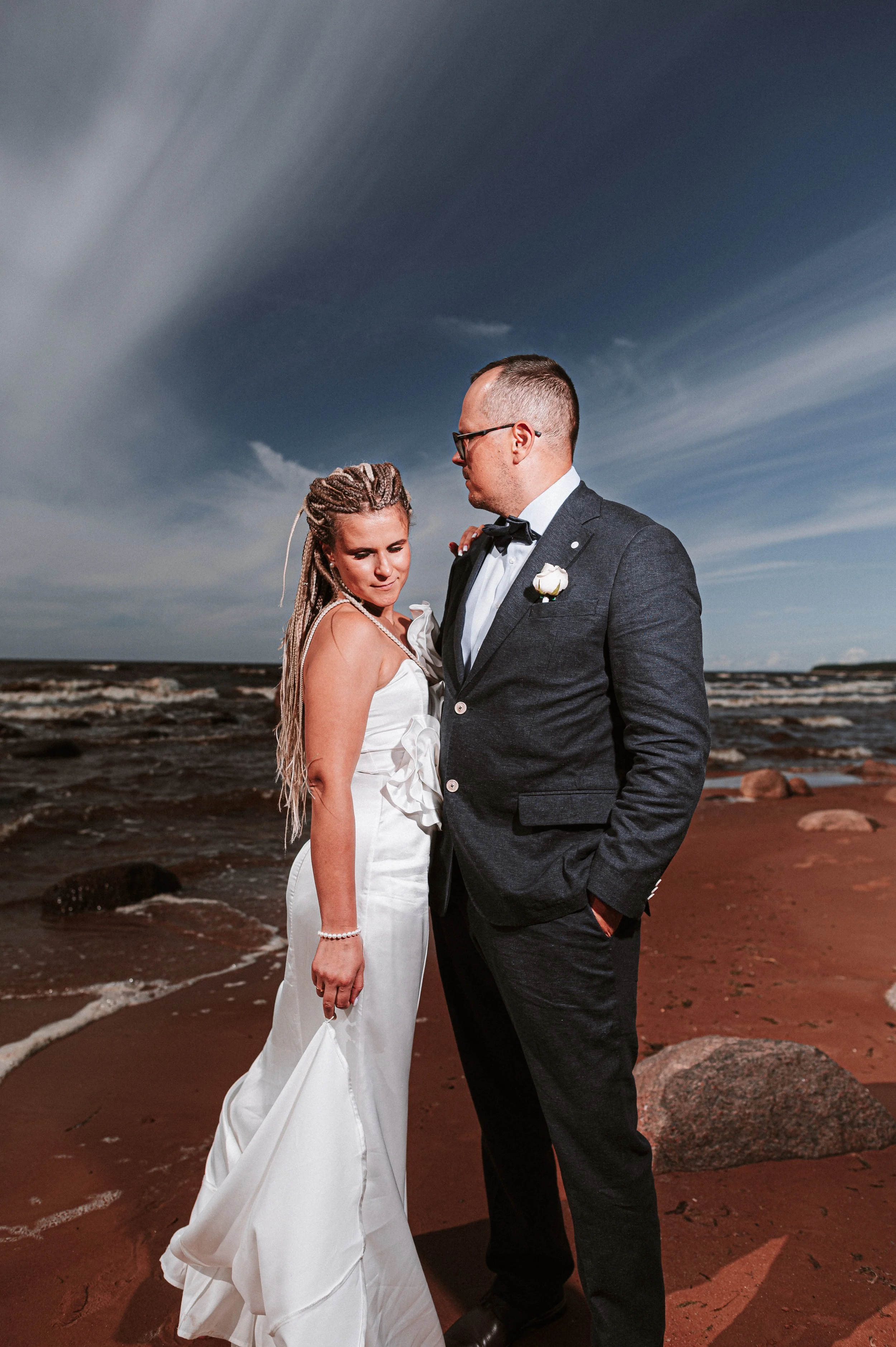 A bride and groom standing on a beach with a cloudy sky in the background. The bride wears a white wedding dress, and the groom wears a dark suit with a bow tie.