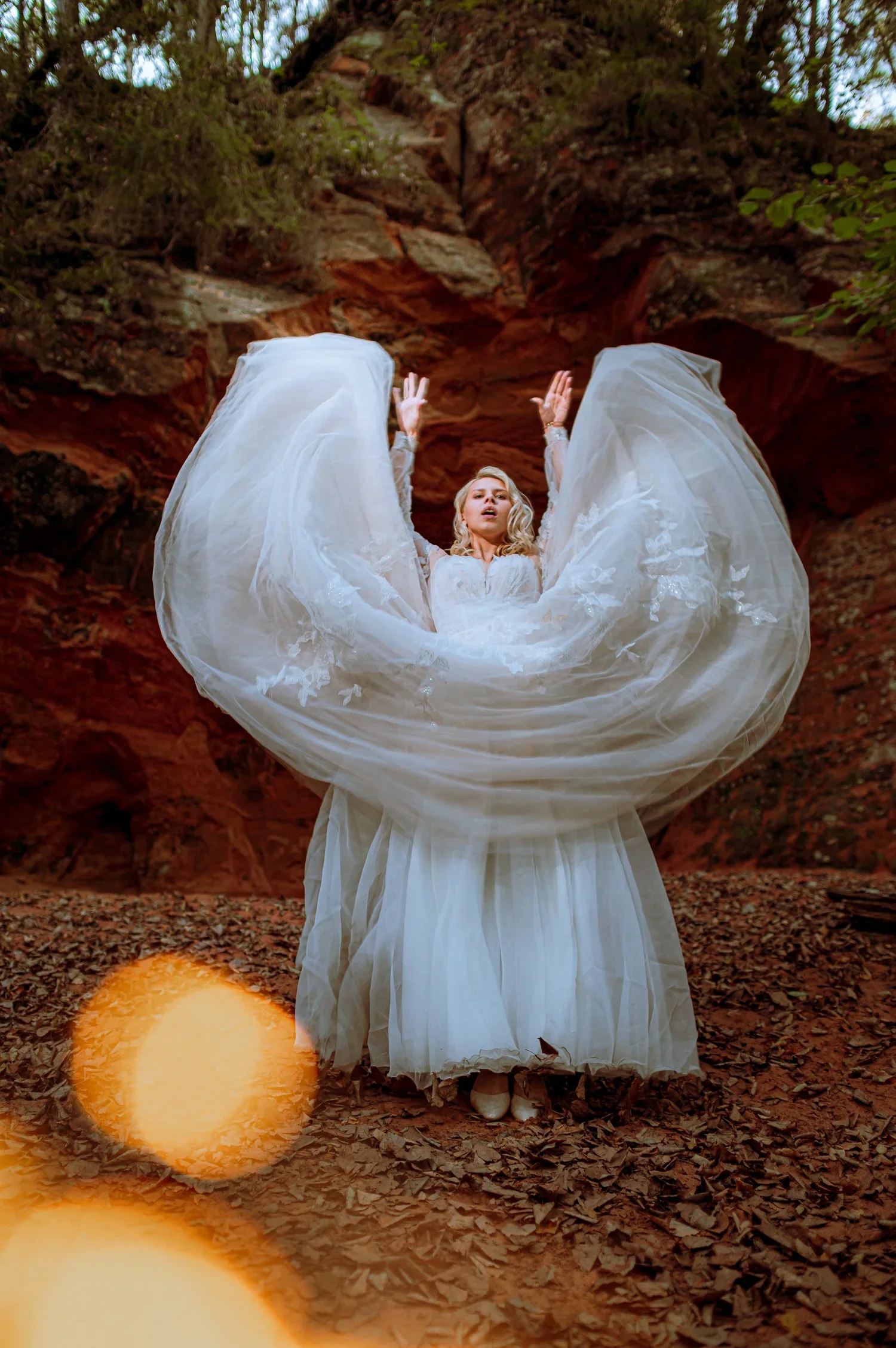 A woman in a white wedding dress standing outdoors against a rocky and wooded background, with her dress billowing around her.