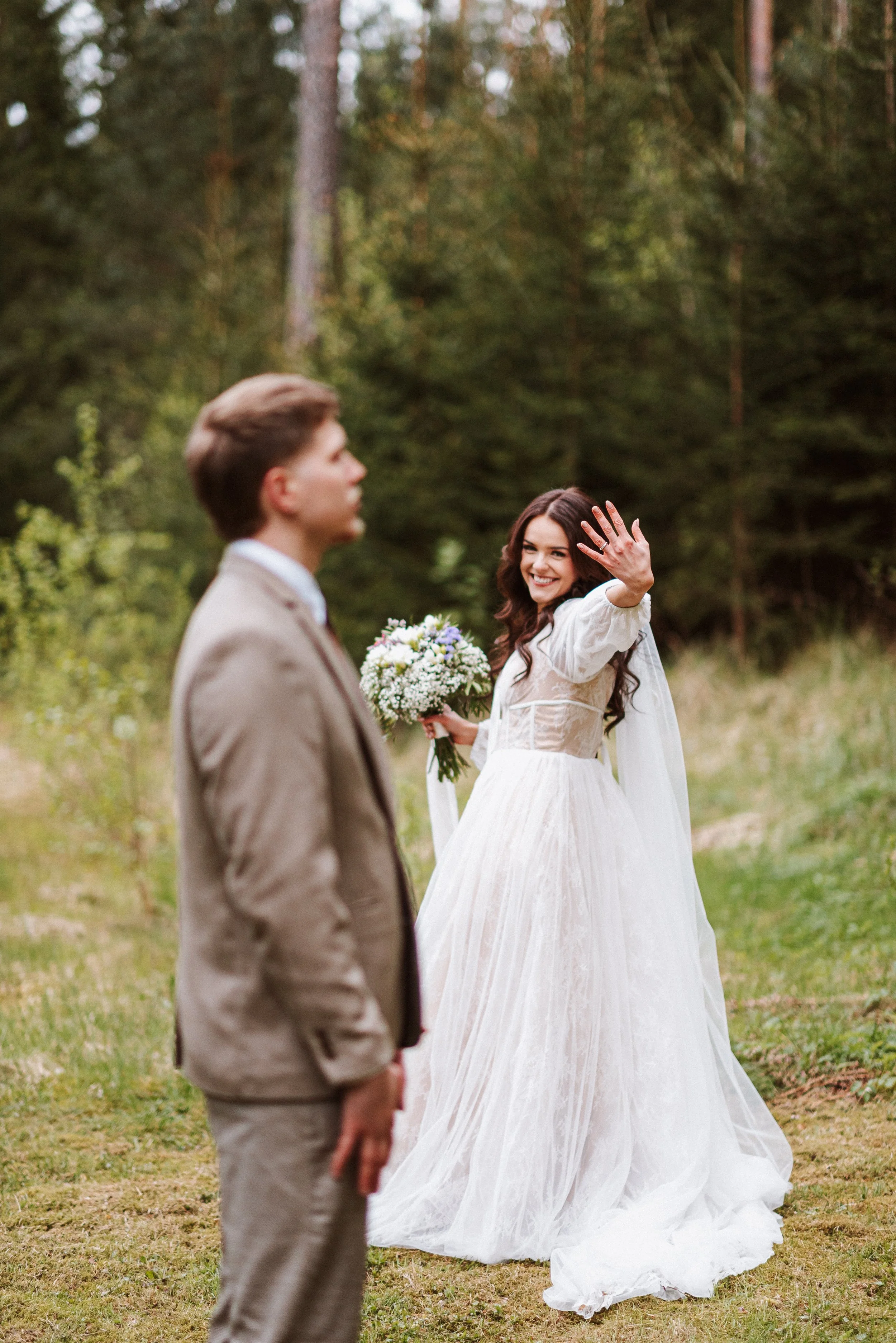 A woman in a white wedding dress happily shows her hand with a wedding ring while holding a bouquet, standing outdoors in a forested area, with a man in a beige suit in the foreground.