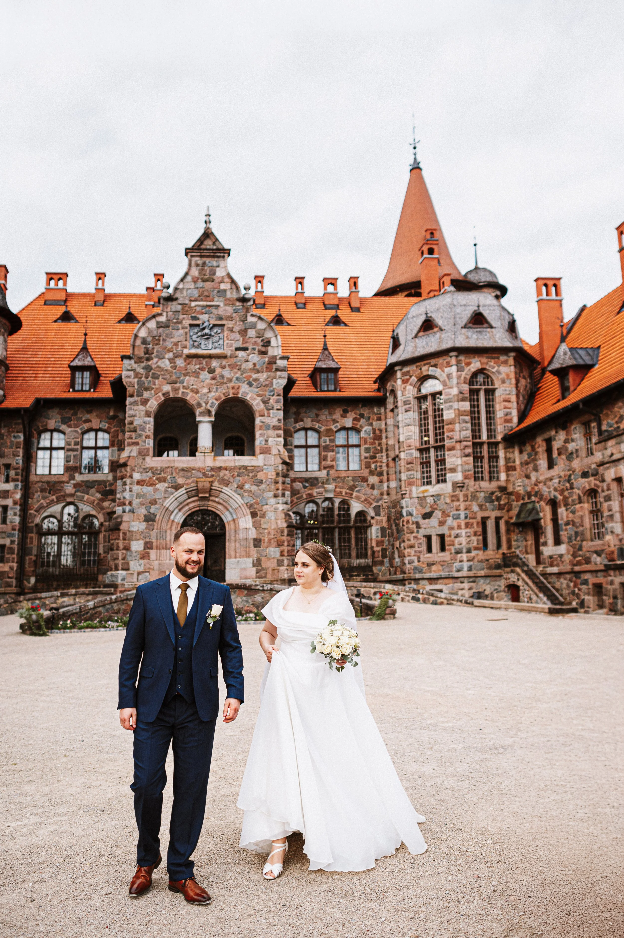 A bride and groom walking outside a historic stone castle with orange rooftops on a cloudy day.