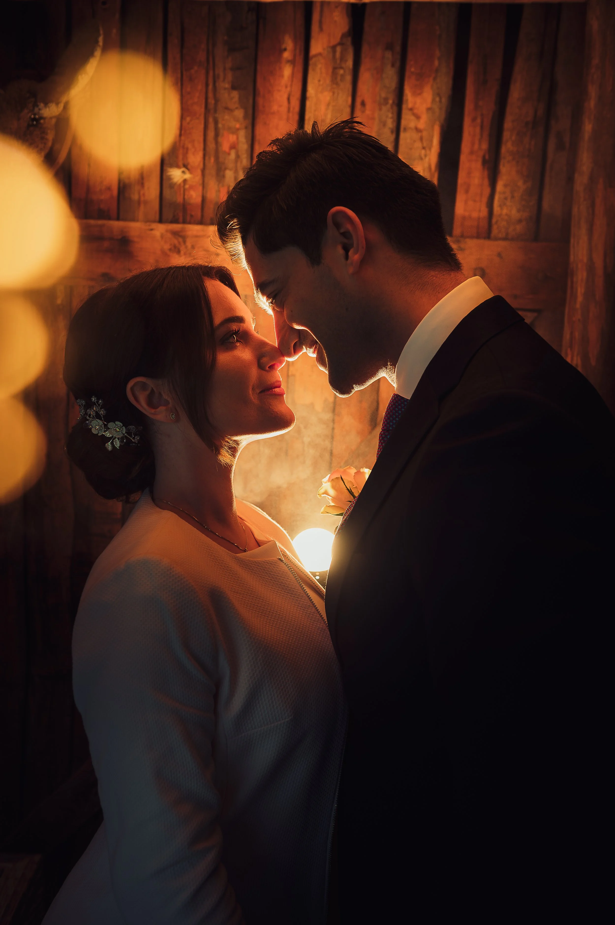 A bride and groom standing close together, facing each other with their foreheads and noses touching during a wedding photo with warm lighting and background wooden wall.