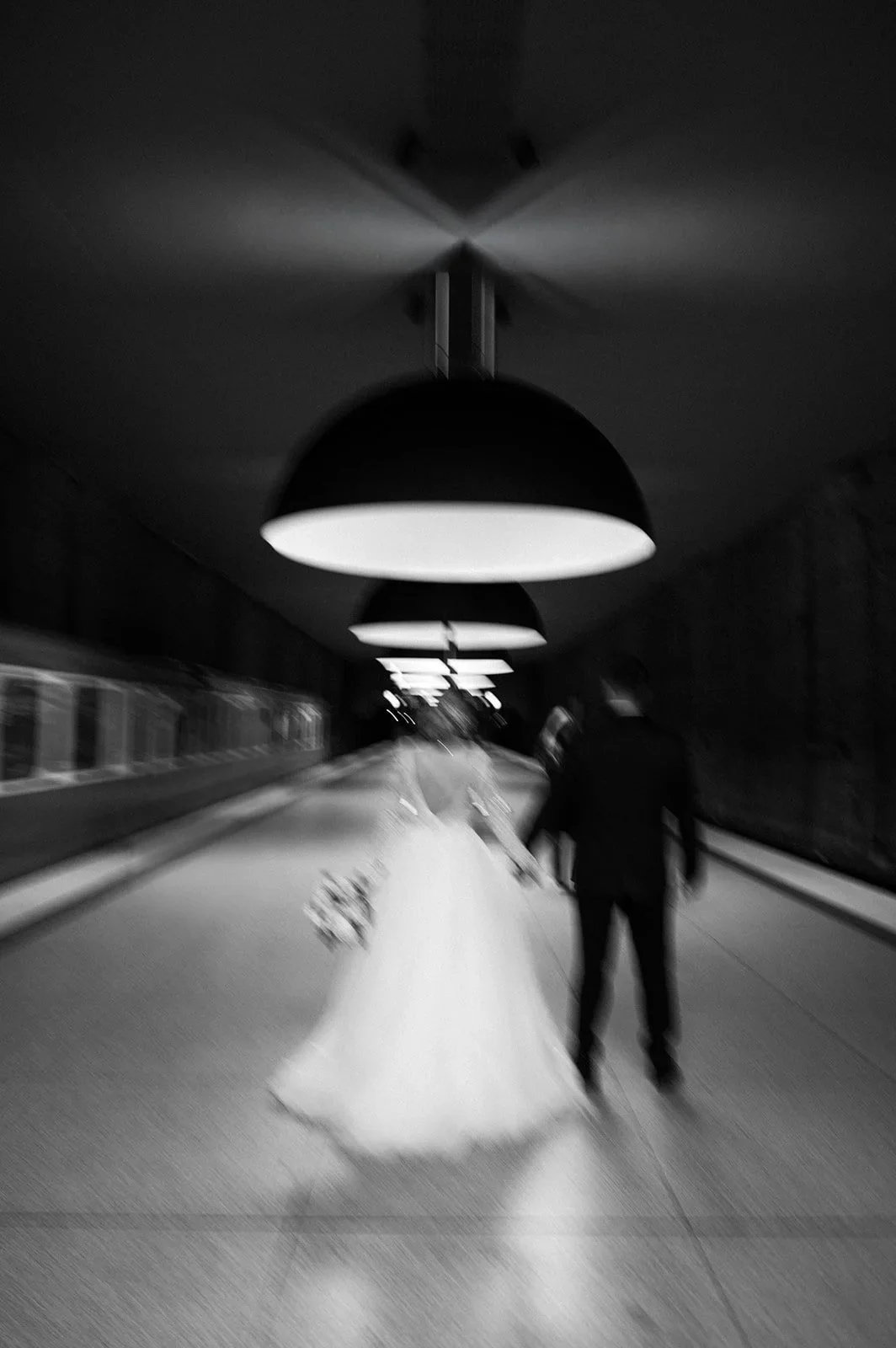 Black and white photo of a bride and groom holding hands and walking away in an underground tunnel, illuminated by row of hanging lights.
