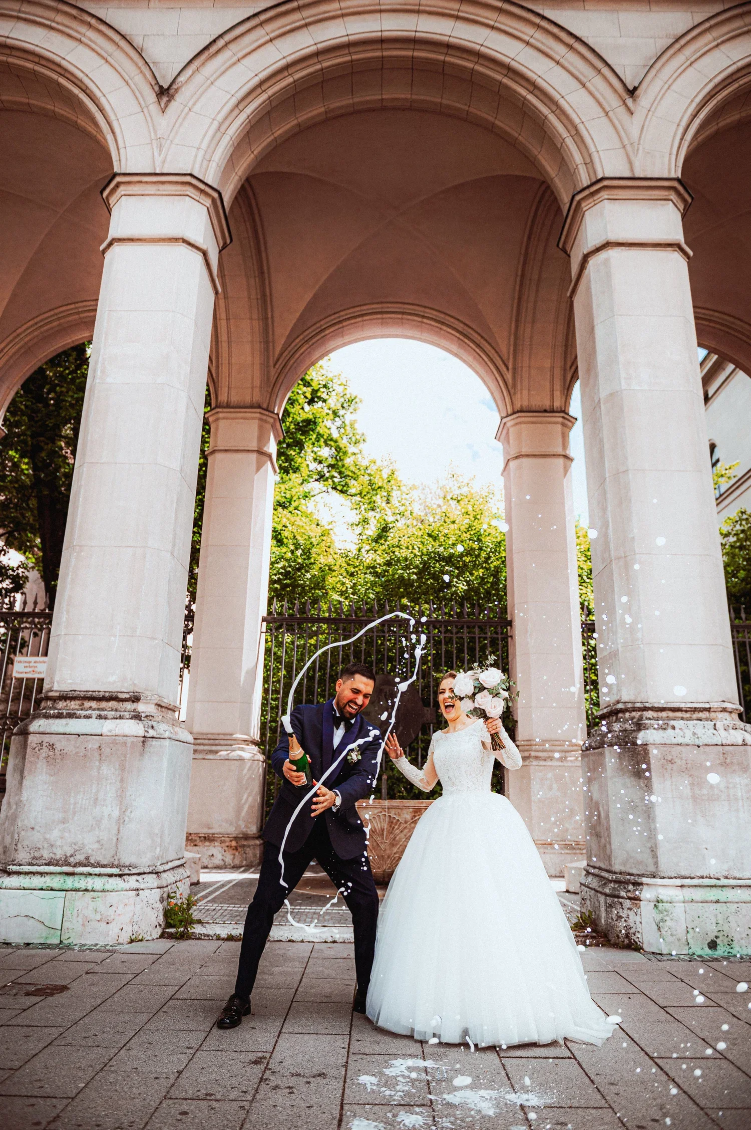 A newlywed couple celebrating outdoors under a stone archway, with champagne and a wedding bouquet, surrounded by celebration spray.