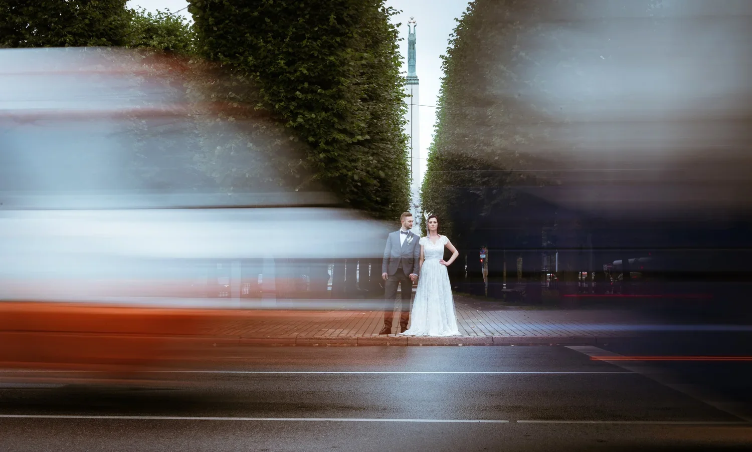 A bride and groom holding hands on a city sidewalk at night, with light trails from passing vehicles and a tall monument visible in the background.