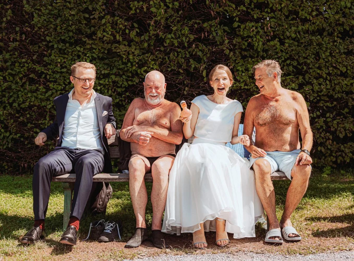 Five people sitting on a park bench laughing, one woman in a white dress, four men shirtless, with two in suits and shorts, against a lush green hedge background.