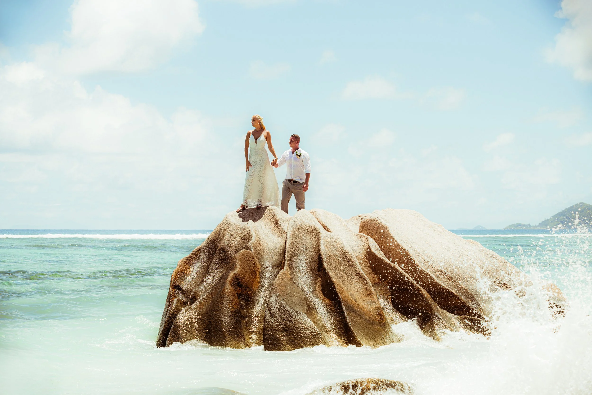 A bride and groom standing on a large rock in the ocean, holding hands, with the sea and sky in the background.
