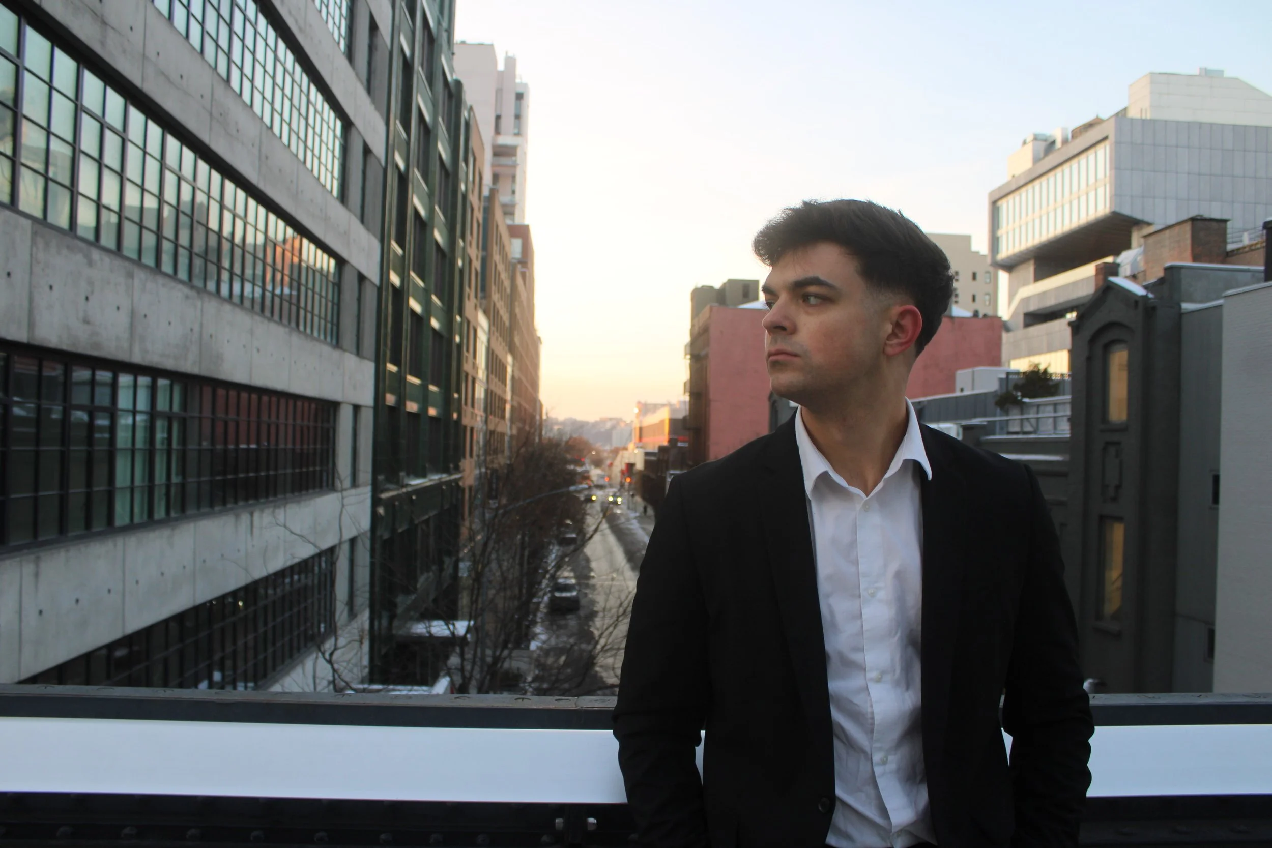 A young man in a black jacket and white shirt standing on a balcony in an urban setting during sunset, with tall buildings and a street below.