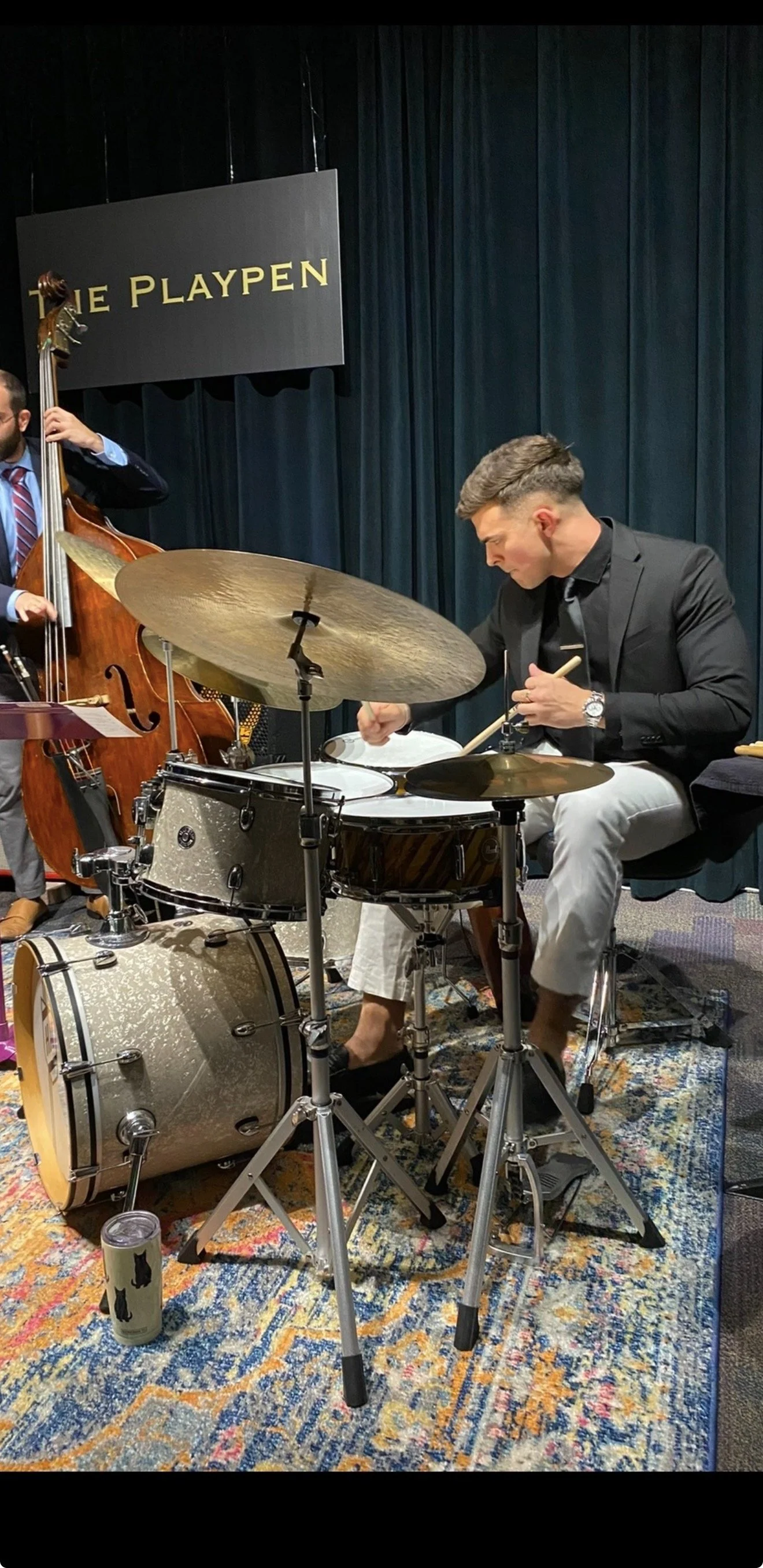 A musician playing drums during a live performance, with a double bass player in the background, on stage with dark blue curtains and a sign that reads 'THE PLAYPEN'.