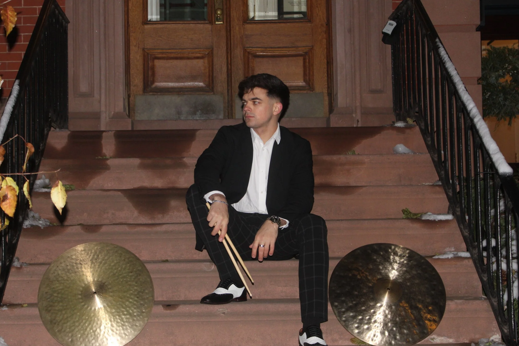 A man sitting on outdoor steps with musical cymbals and drumsticks, wearing a black blazer, white shirt, plaid pants, and dress shoes, near a brown wooden door.