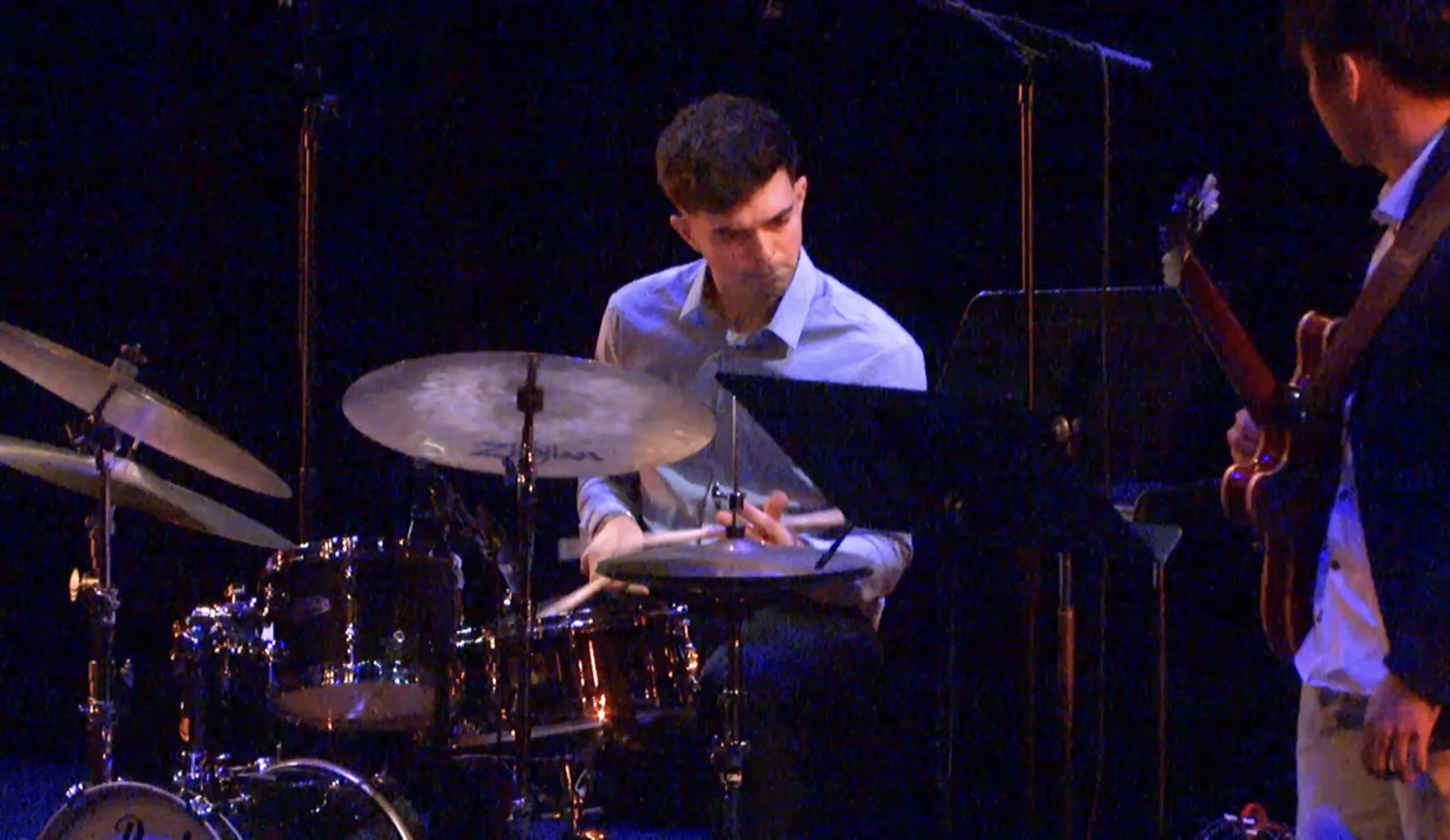 Two musicians performing on stage, one playing drums and the other playing a bass guitar, with a dark background.