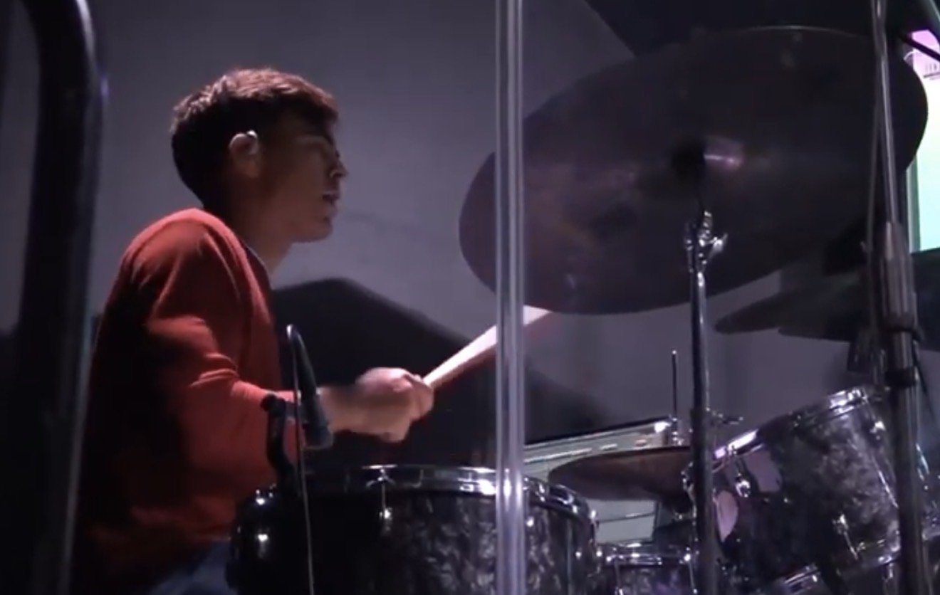 A young man playing the drums on stage, wearing a red shirt, with dark studio lighting.