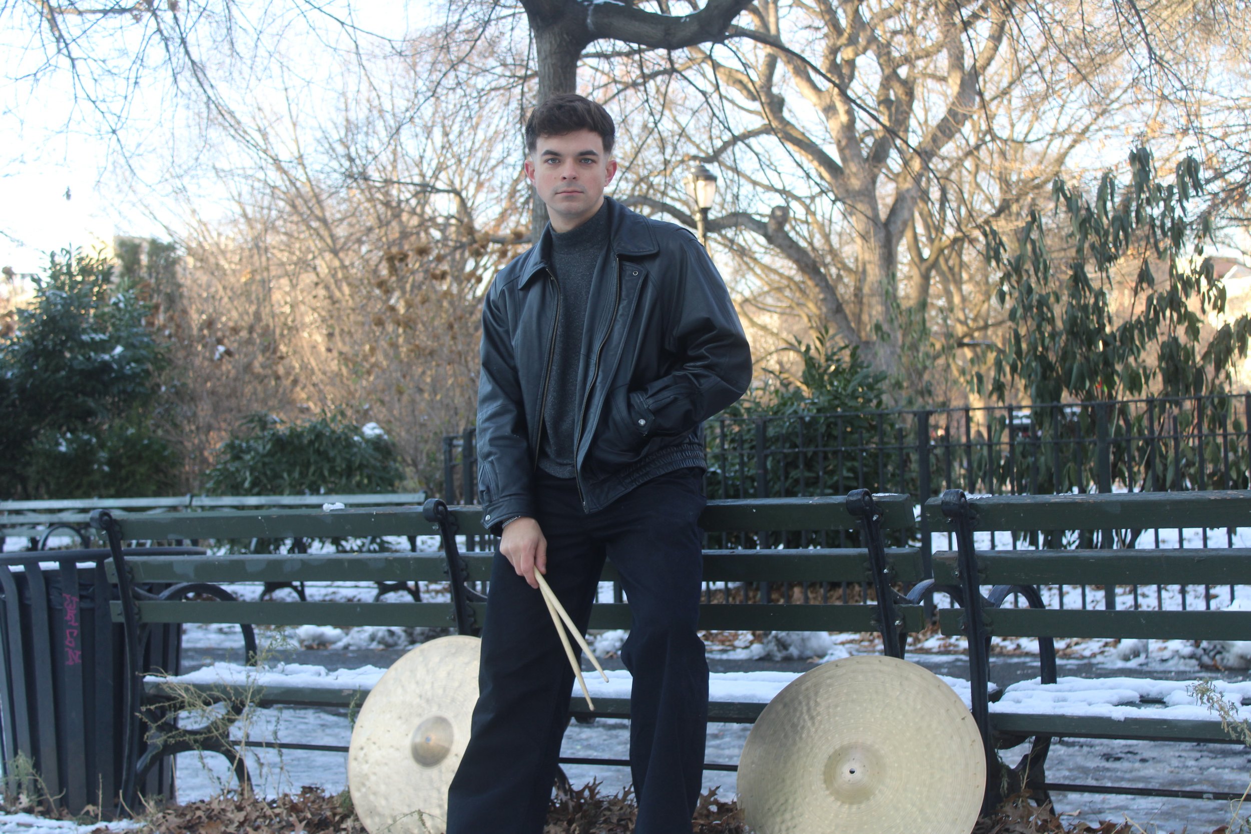 A young man with dark hair standing in a park with snow, holding drumsticks, with two large cymbals on the ground, wearing a black leather jacket, dark sweater, and black pants, with leafless trees and benches in the background.