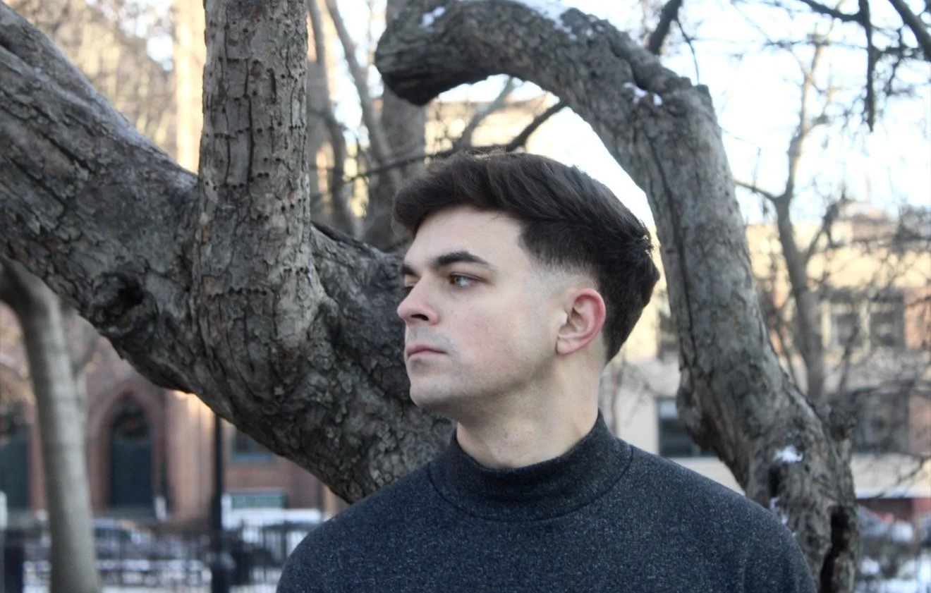 A young man with dark hair and a side undercut stands outdoors near a leafless tree, looking to the side with a serious expression.