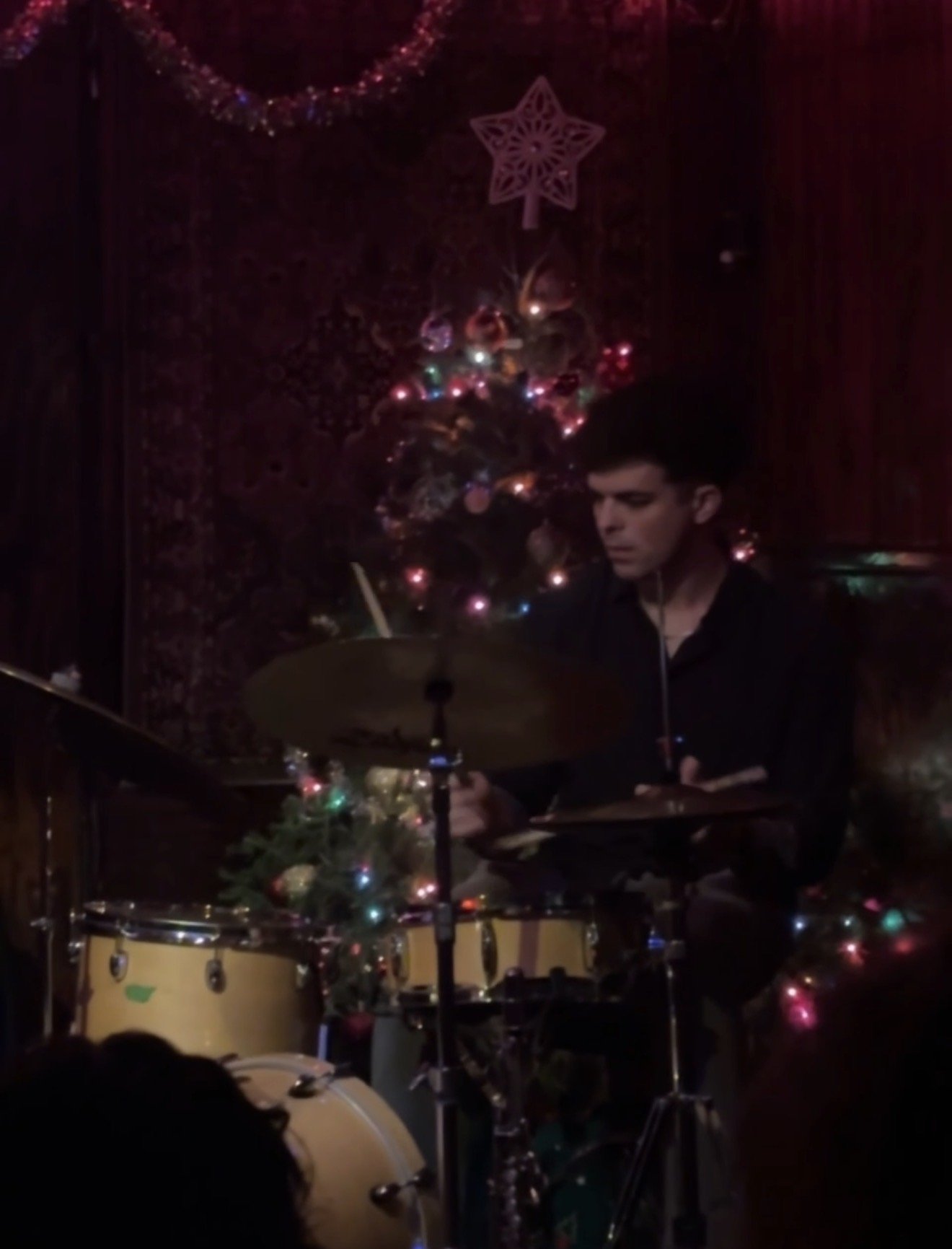 A person playing drums on a Christmas decorated stage with a small Christmas tree and ornaments, in a dimly lit room.