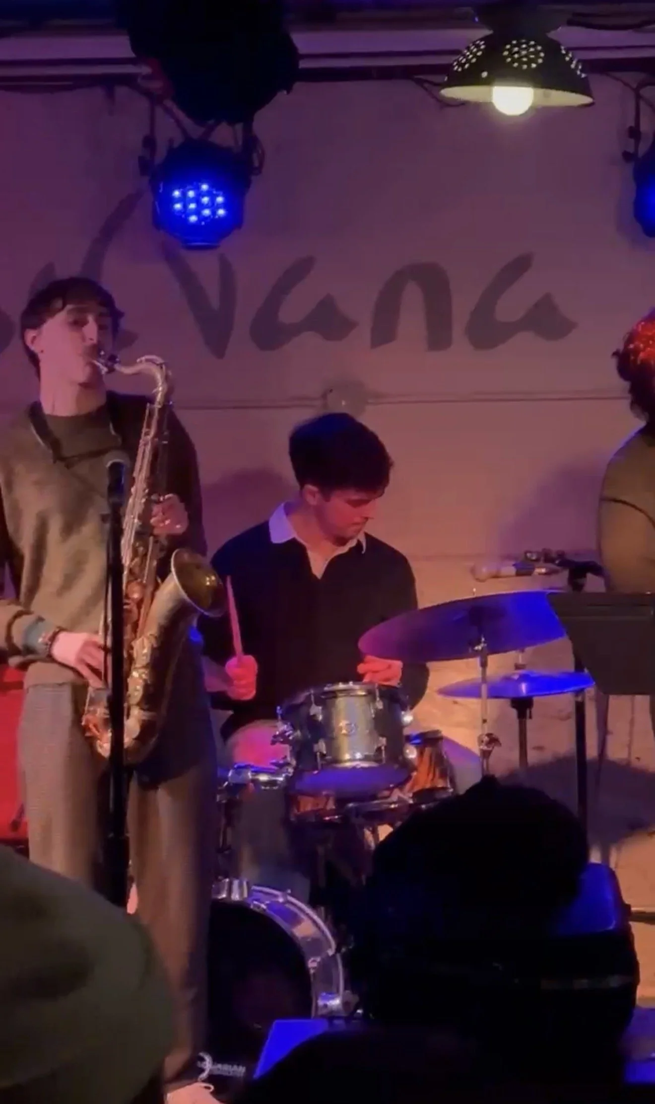 Musicians performing live on stage with a saxophonist and drummer, under purple and blue stage lights. The background reads 'Vana'.