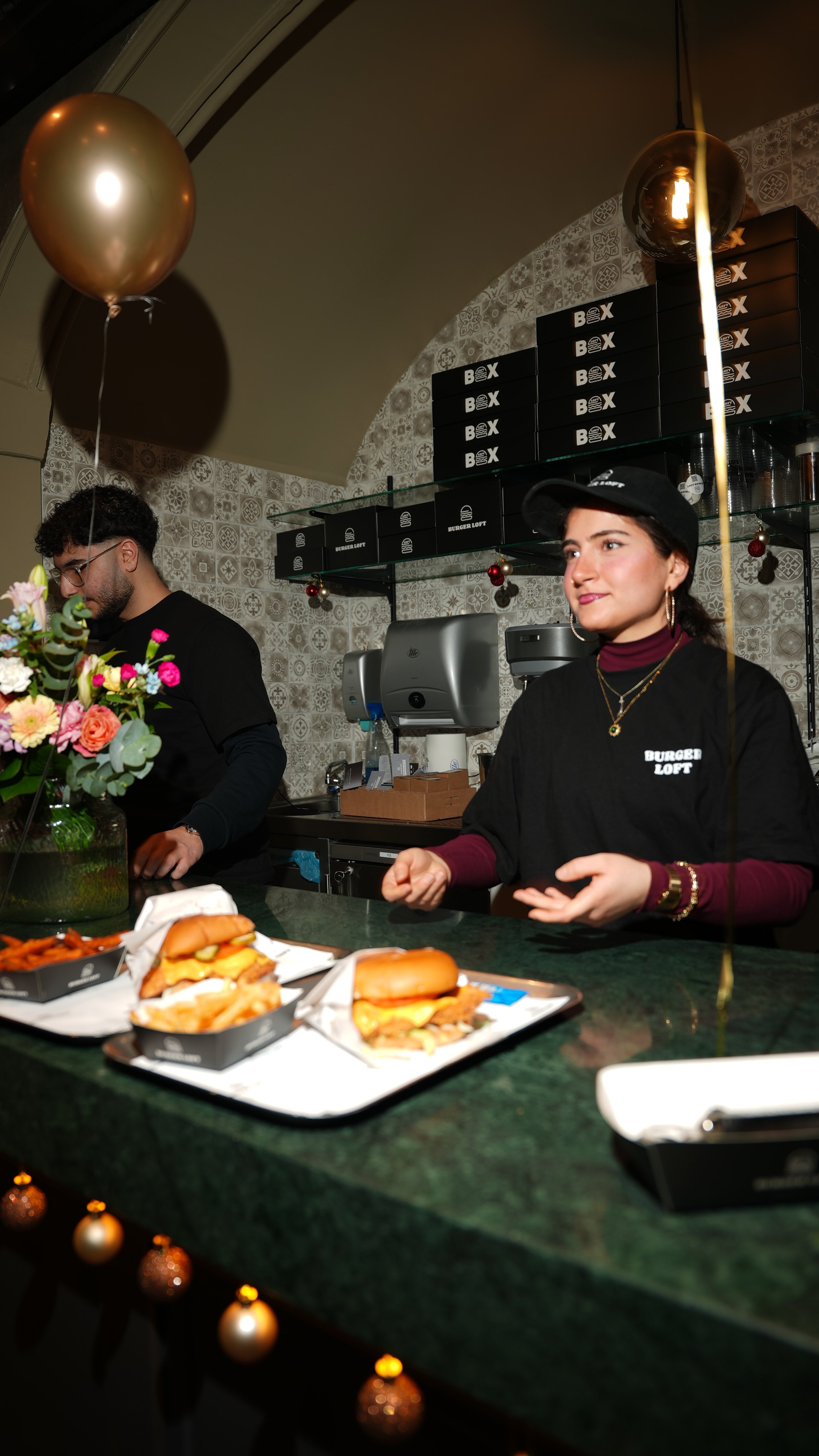 Zwei Mitarbeiter in einem Imbissrestaurant hinter der Theke, auf dem Tisch liegen Fast-Food-Burger, Pommes und andere Snacks, im Hintergrund gibt es Schwarz-Weiß-Kunst auf der Wand, Weihnachtsdekoration und eine goldene Ballon.