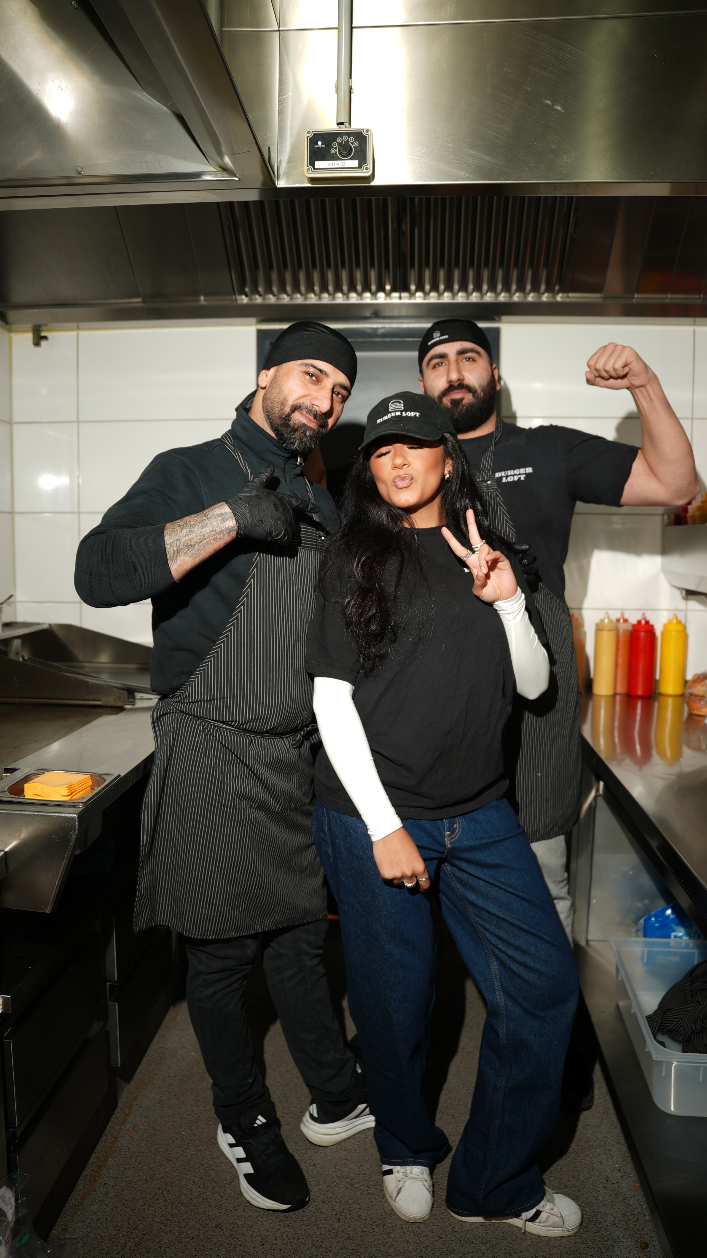 Drei Köche und eine Frau in einer Restaurantküche, die posieren und winken, mit verschiedenen Handzeichen. Sie tragen schwarze Kleidung und Schürzen.