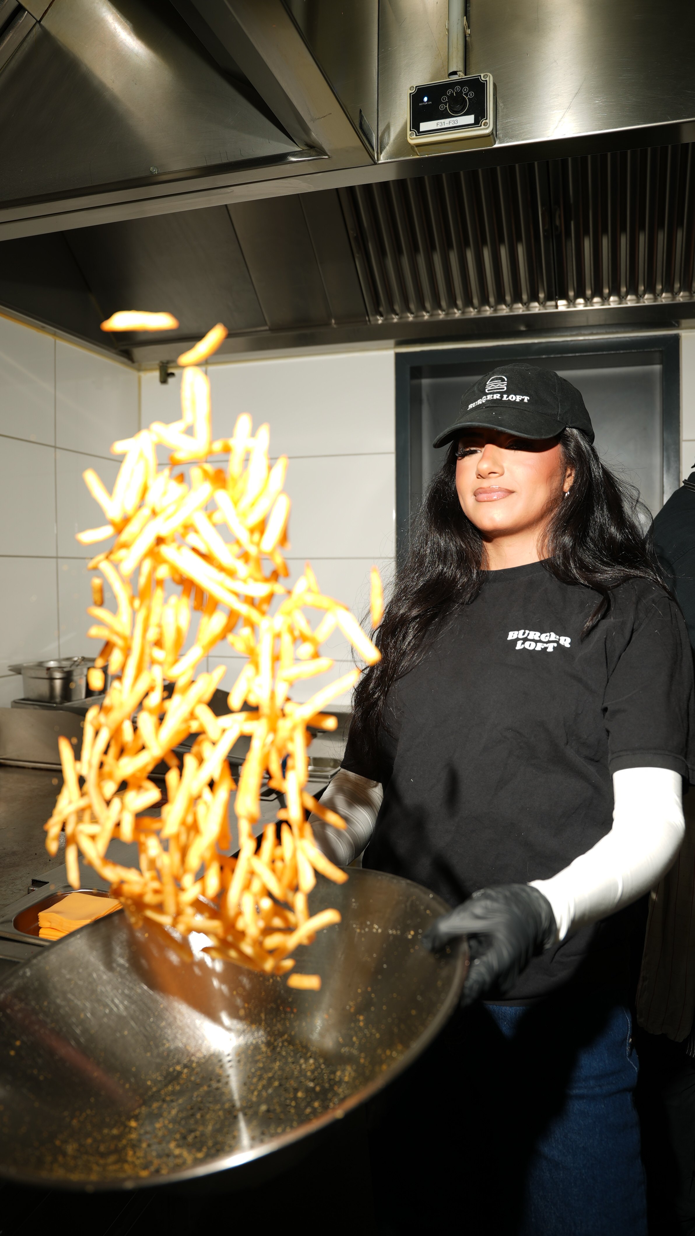 Eine Frau in Schwarz mit einer schwarzen Cap und schwarzen T-Shirt beim Frittieren von Pommes in einer Küche.
