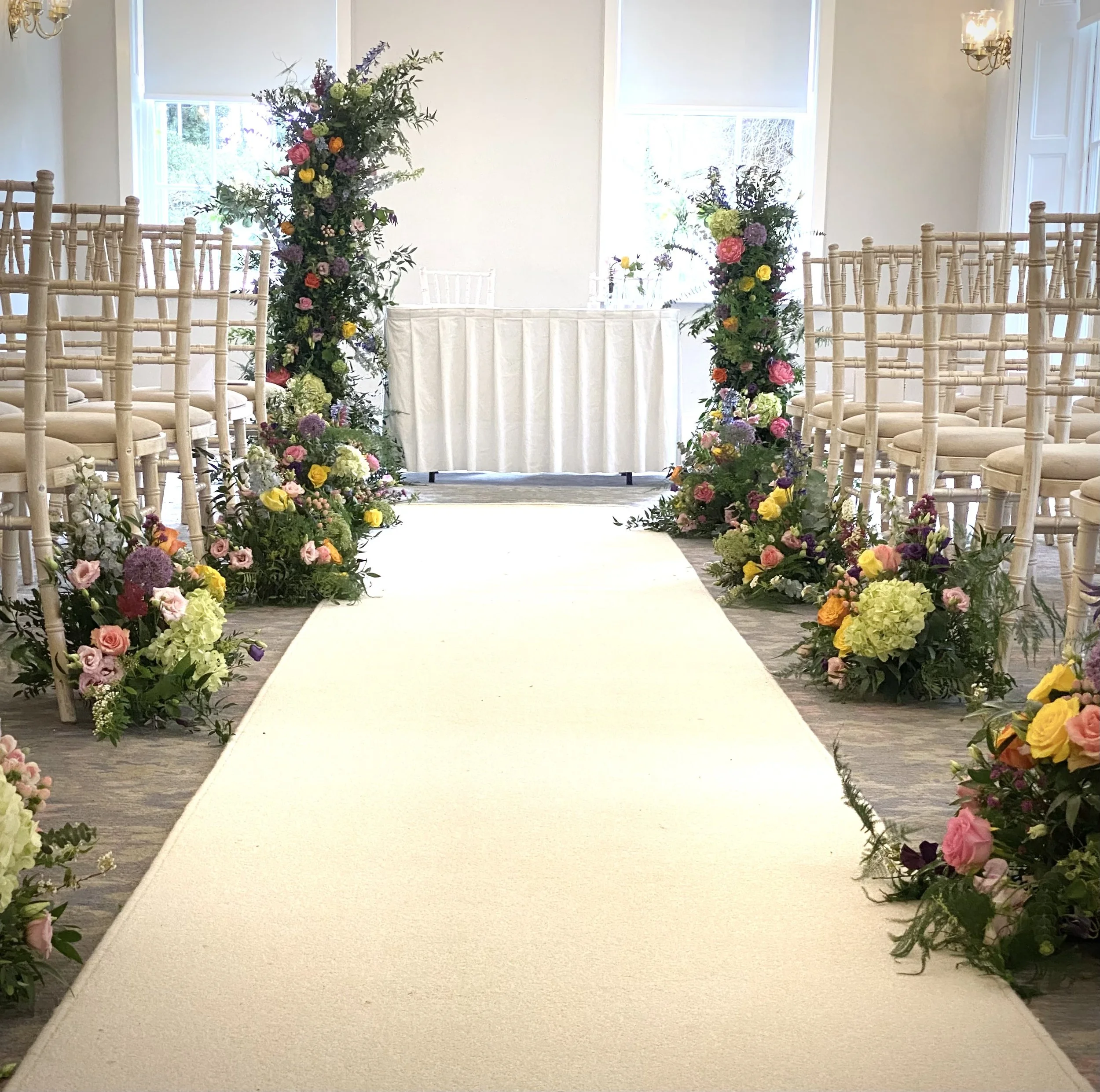 Indoor wedding ceremony setup with floral arches on either side of a white aisle rug, beige chairs arranged on both sides, and sunlight coming through windows.