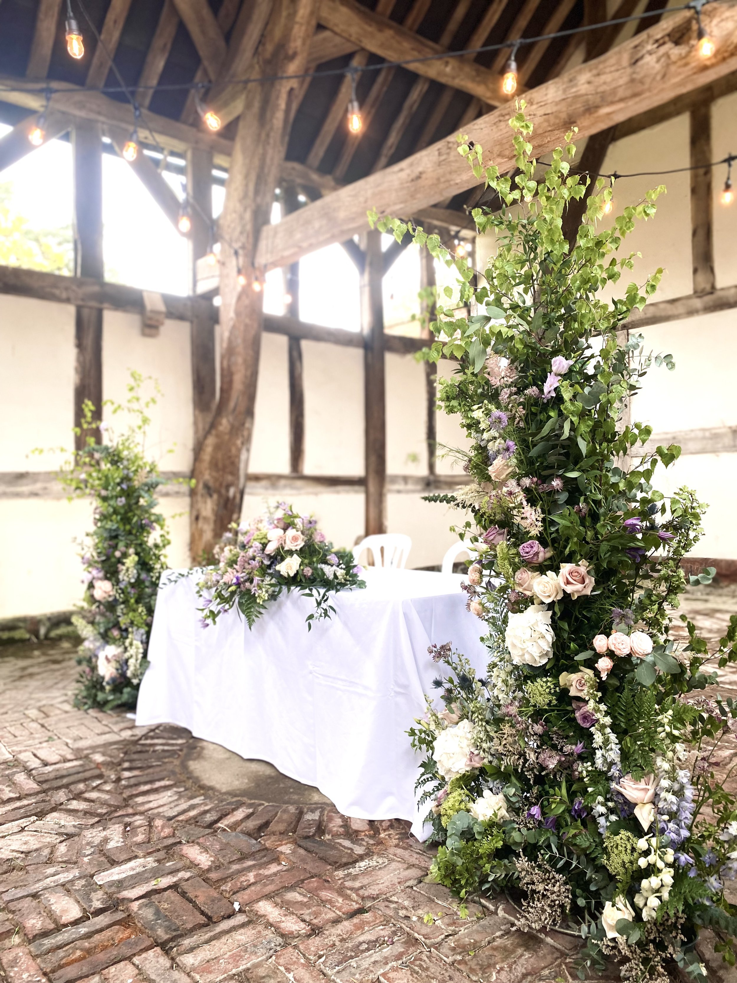 Wedding reception table decorated with floral arrangements under a rustic wooden barn ceiling with string lights.