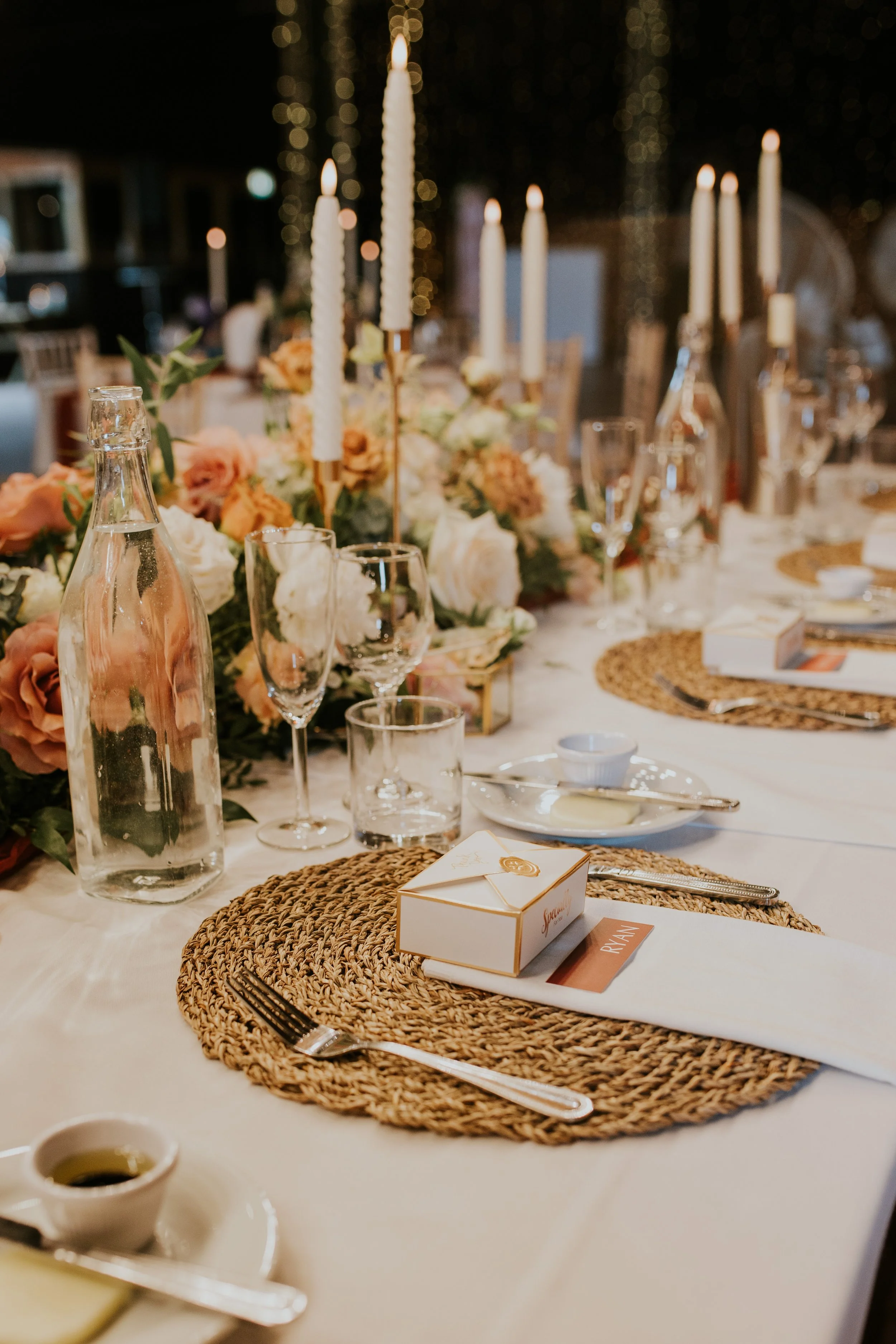 Elegant banquet table set with woven placemats, glassware, candles, floral centerpieces, and place cards for a wedding reception.