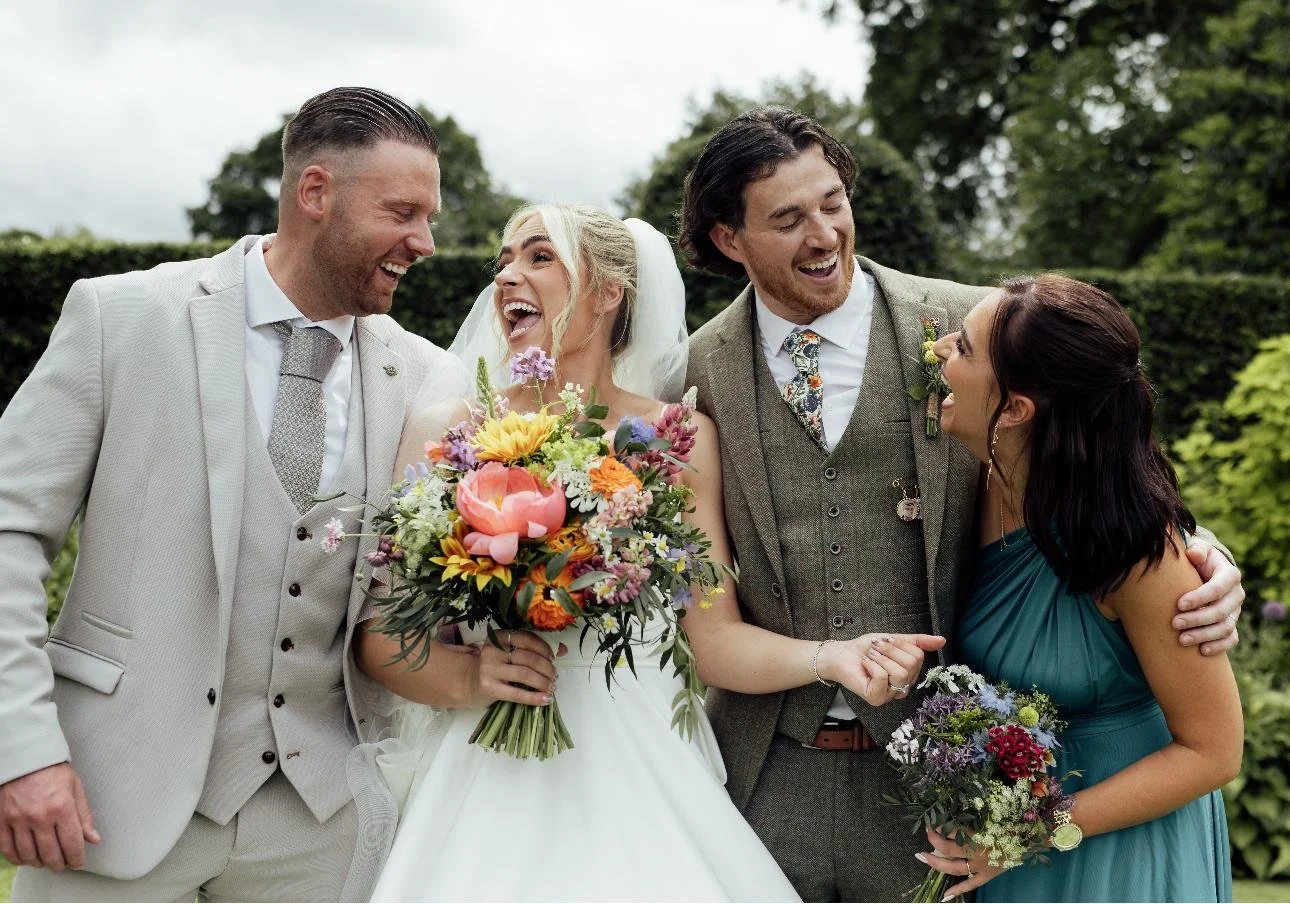 Group of four people at a wedding, including the bride in a white dress holding a bouquet, and three others dressed in formal attire, standing outdoors with green trees in the background.