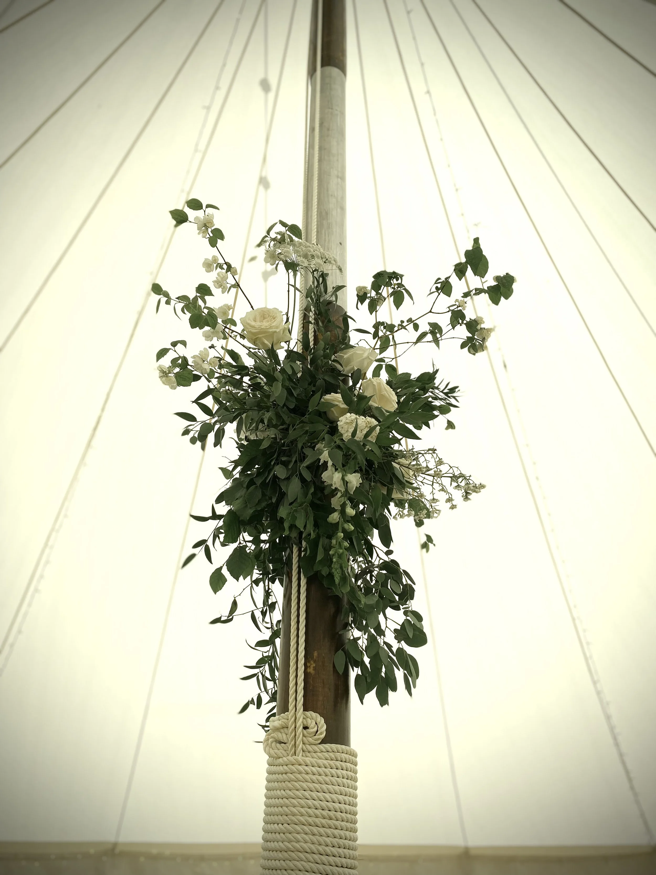 A floral decoration with white roses and green foliage attached to a central pole inside a tent or canopy.