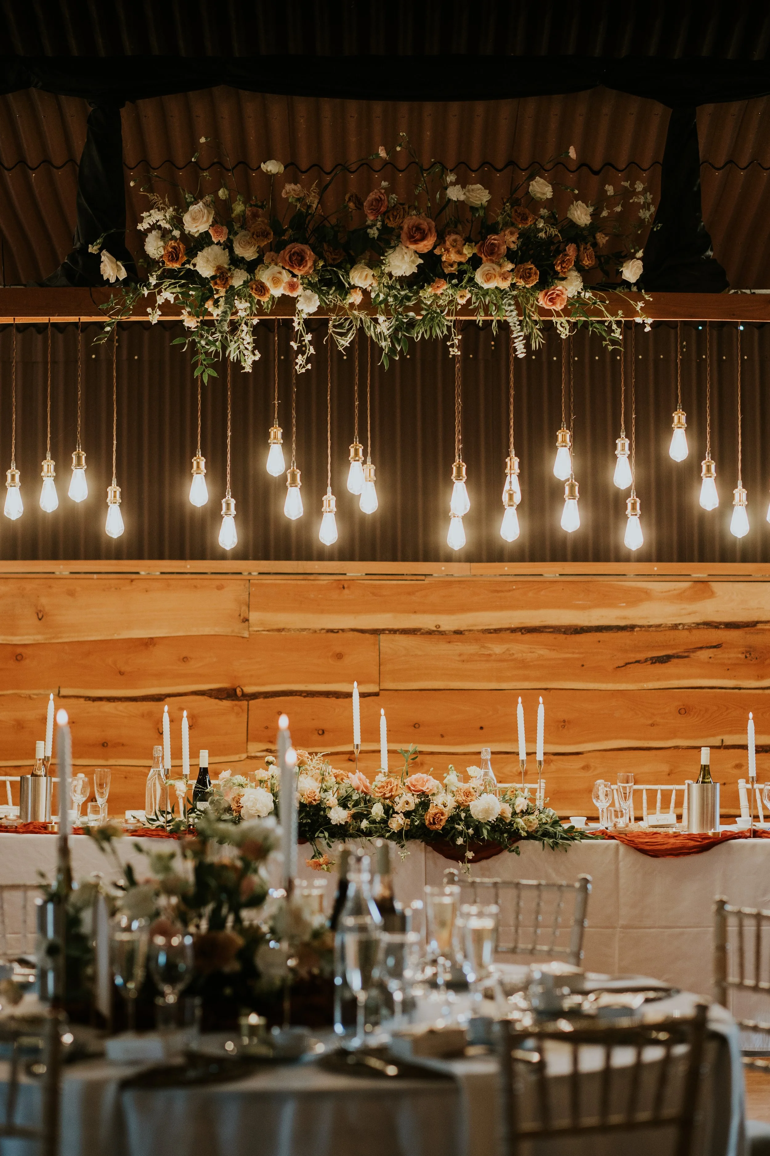 Elegant wedding reception table with candles and floral arrangements, hanging Edison bulbs, and a wooden wall backdrop.