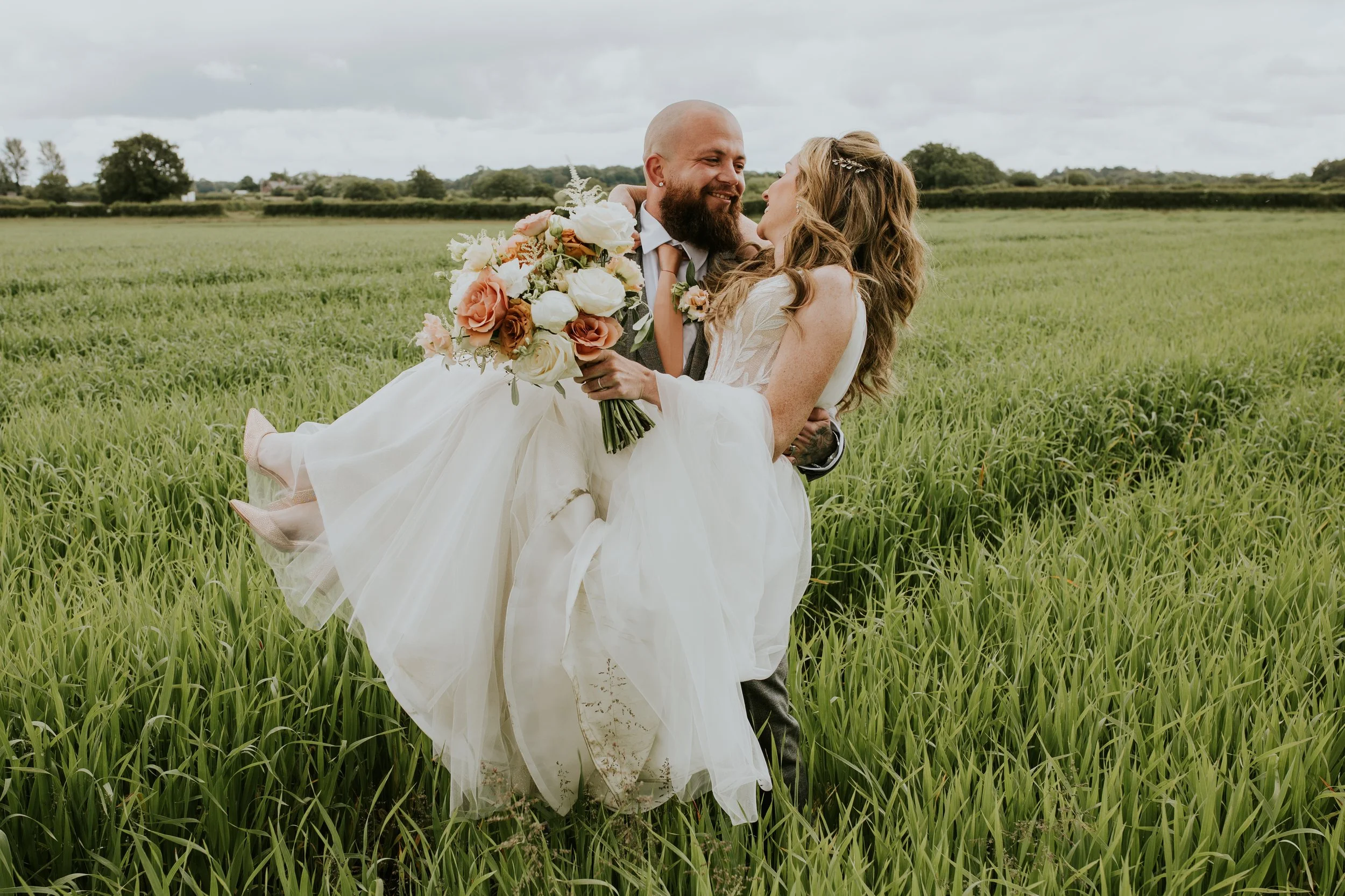 A couple dressed in wedding attire, with the groom carrying the bride, standing in a green field with cloudy sky overhead.
