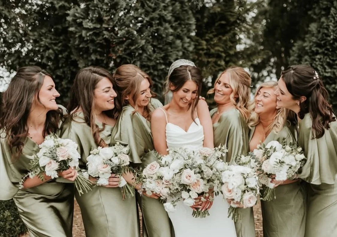 Bride in white dress with her bridesmaids in olive green dresses, all holding bouquets of white and blush pink flowers, outdoors with trees in the background.