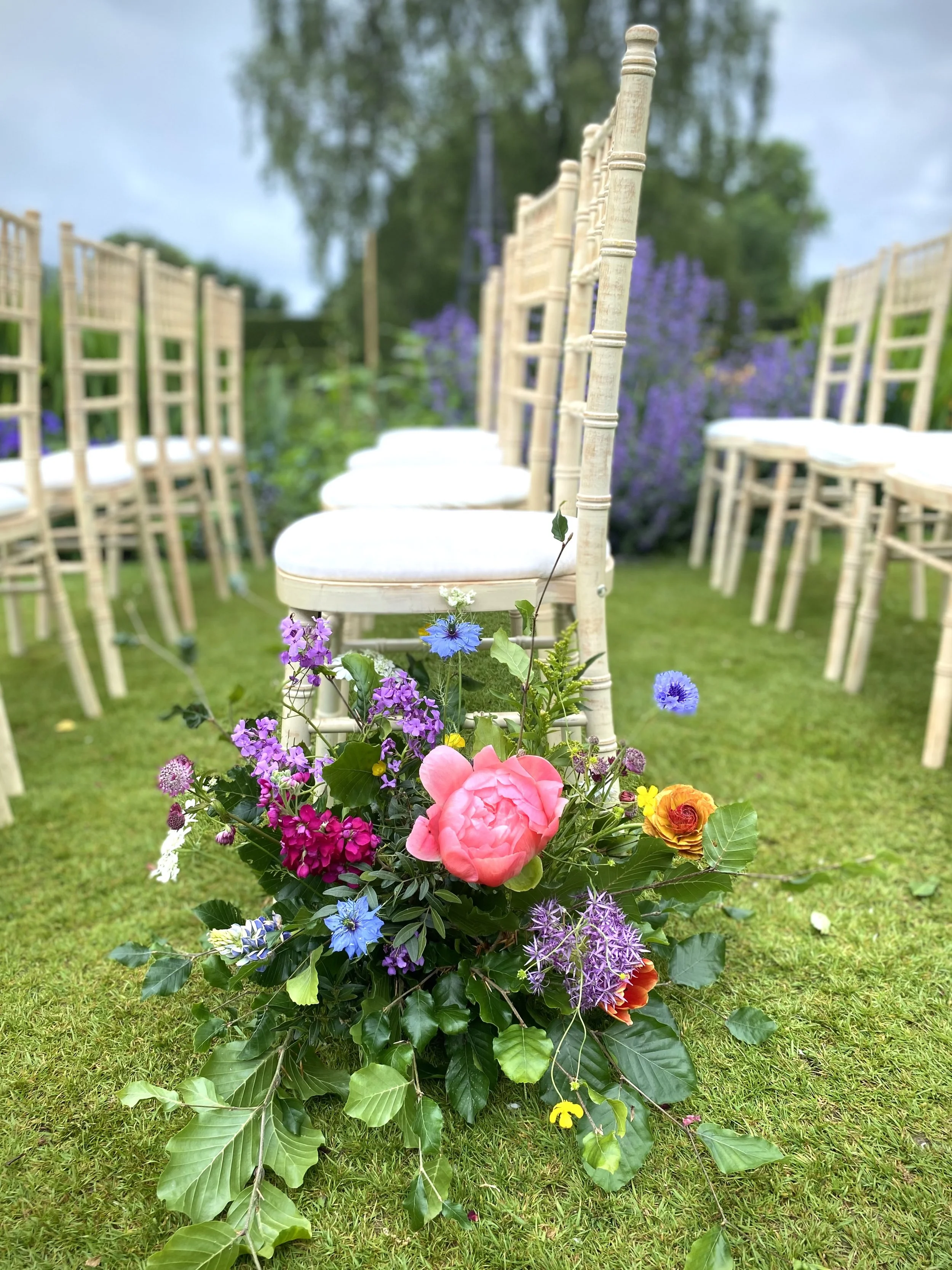 Flower arrangement with pink, purple, blue, yellow, and orange flowers placed on green grass, with outdoor chairs in the background.