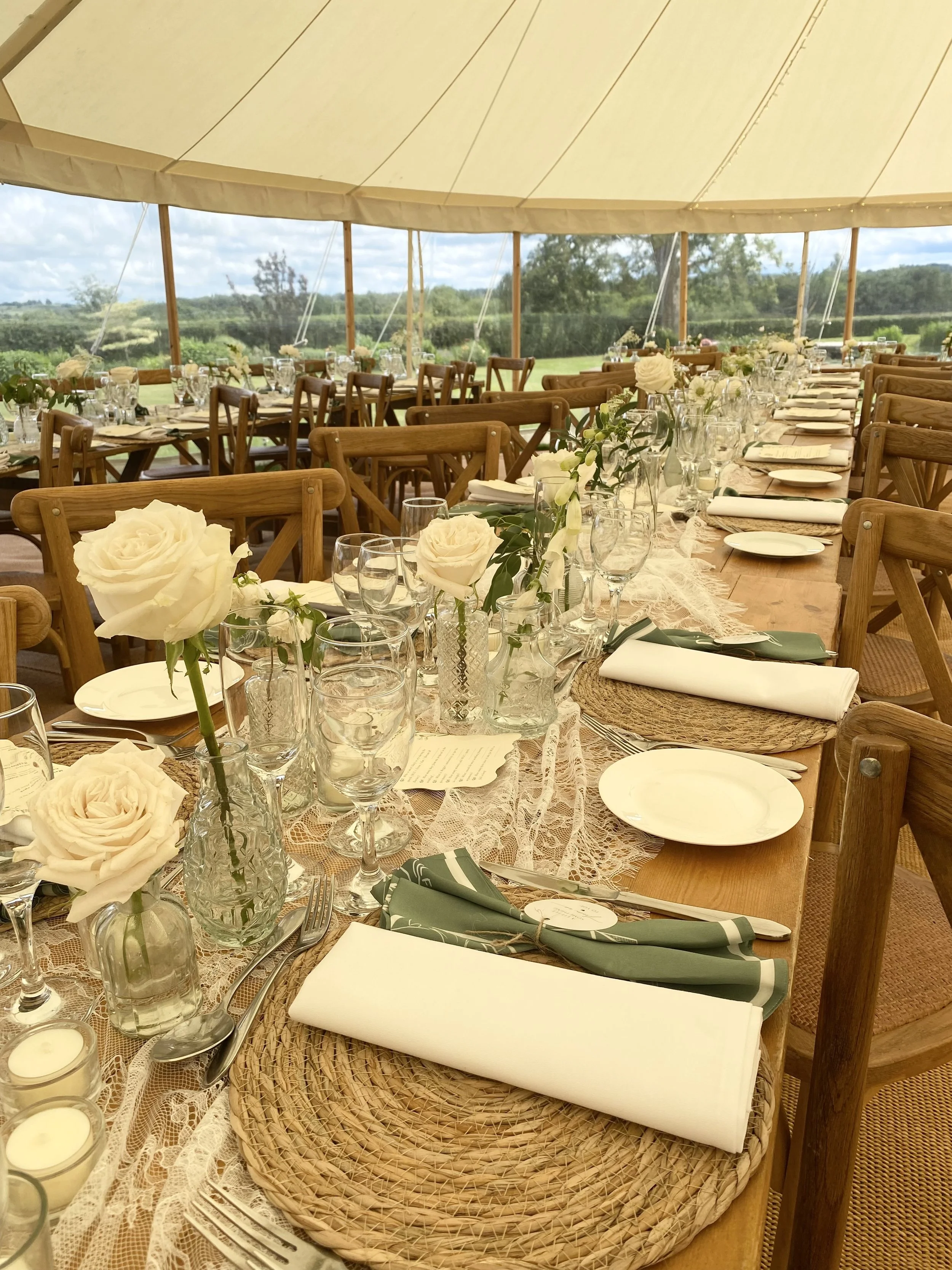 Long banquet table set with floral centerpieces, candles, white plates, green napkins, and glassware under a tent with a scenic outdoor view.