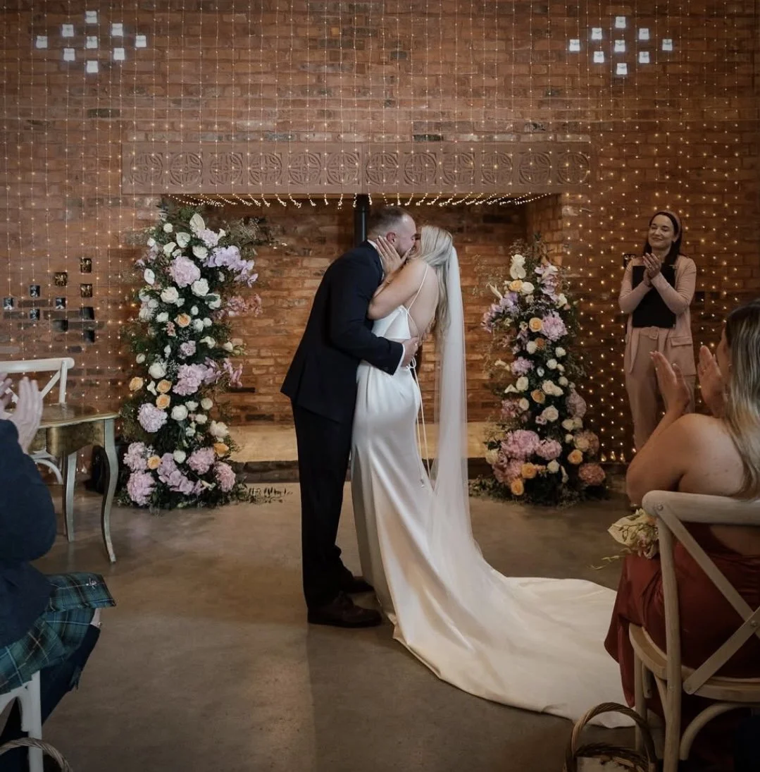 A bride and groom share a kiss at their wedding ceremony. They are surrounded by floral arrangements and decorative lighting on a brick wall. Guests are clapping and watching.