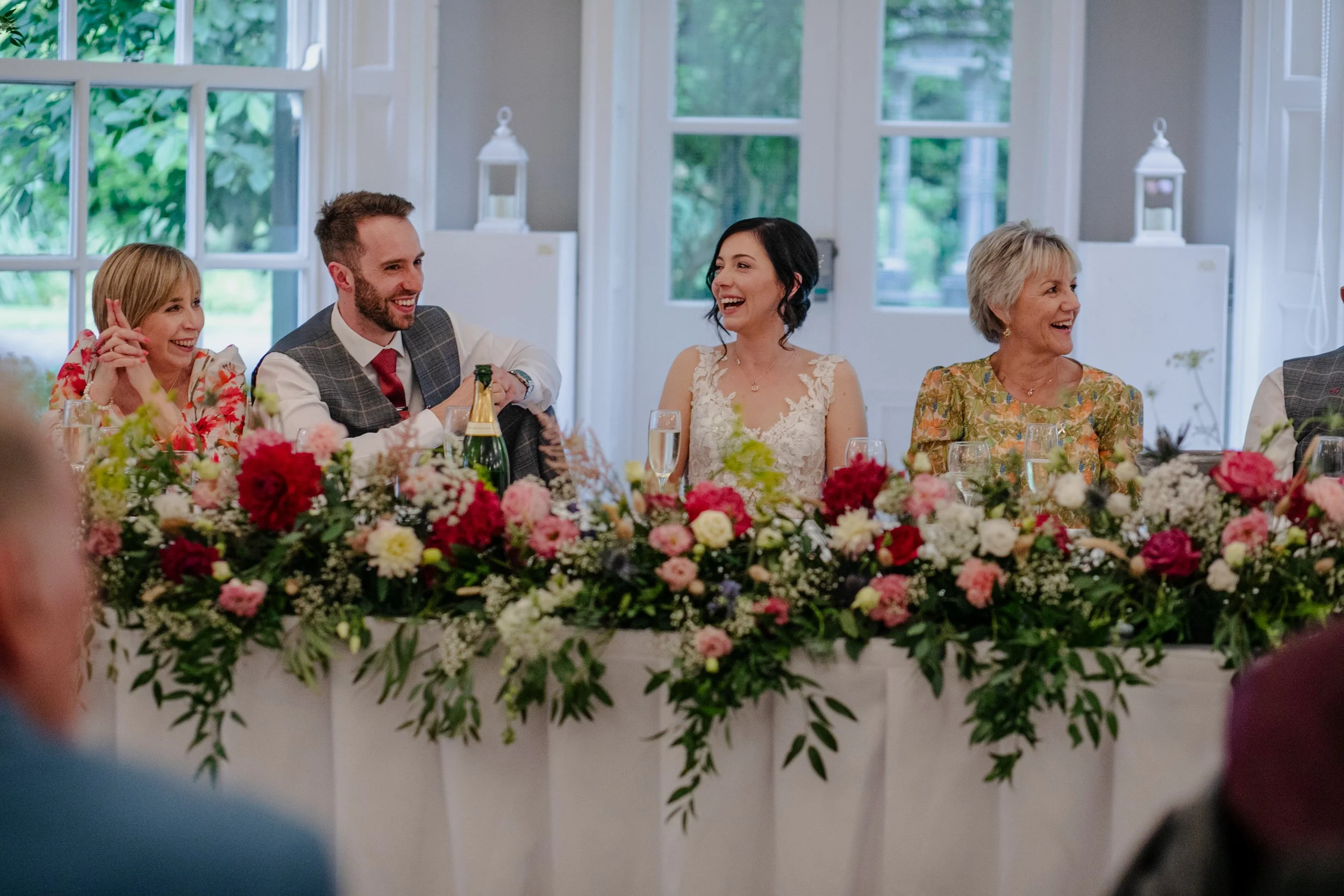 People sitting at a wedding reception table decorated with flowers, smiling and laughing.
