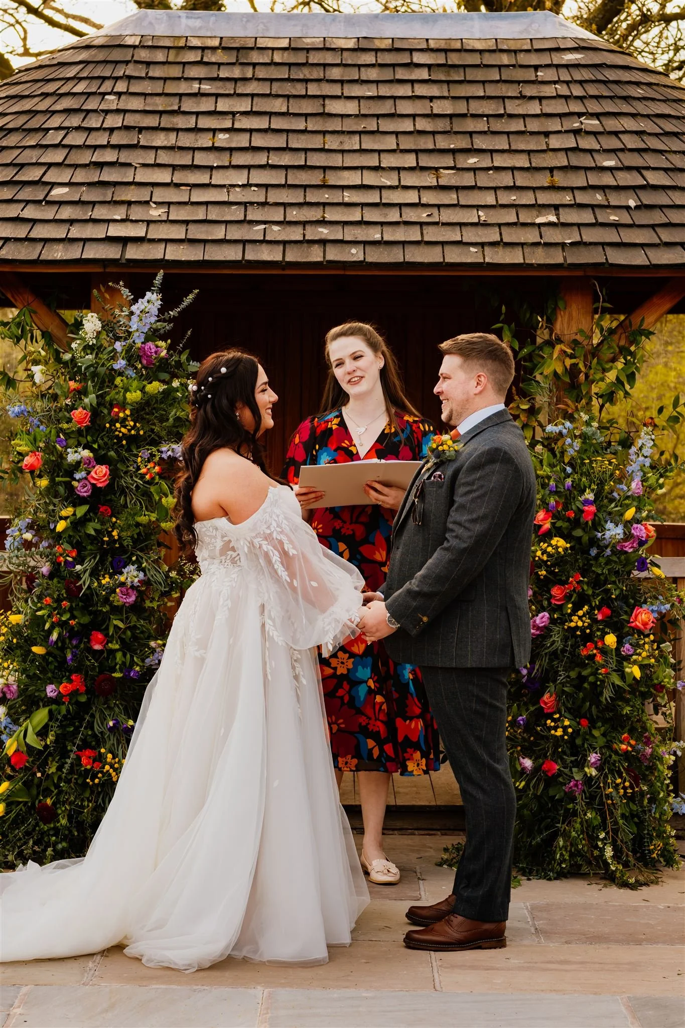 A wedding ceremony with a bride and groom holding hands, exchanging vows, during an outdoor wedding. An officiant stands behind them, reading from a book. The couple is in front of a floral arch with colorful flowers.