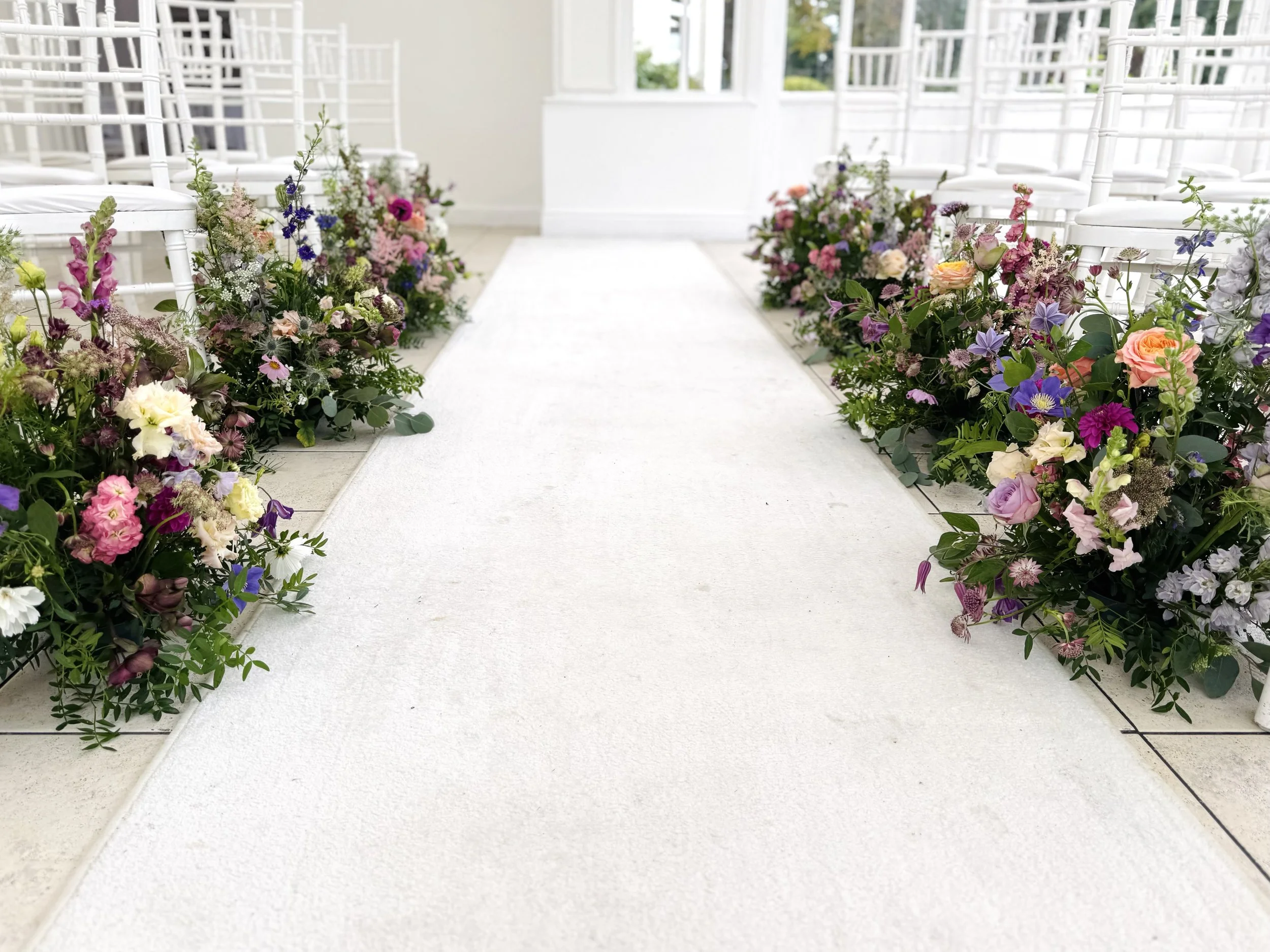 Wedding aisle decorated with colorful flowers on both sides and white chairs.