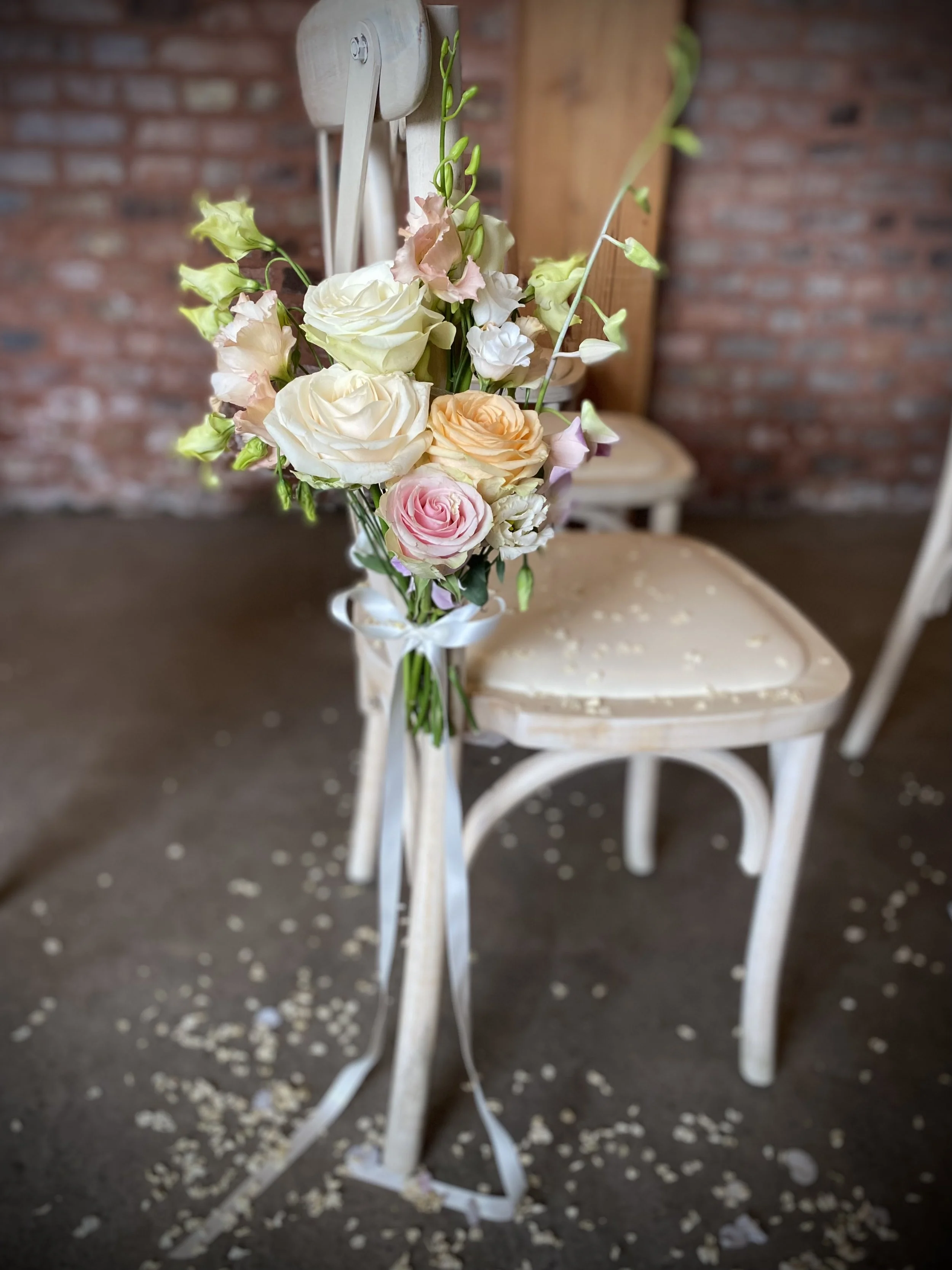 A bouquet of pastel-colored flowers, including roses and lisianthus, tied to a white chair with a ribbon. The background features a brick wall and another white chair.