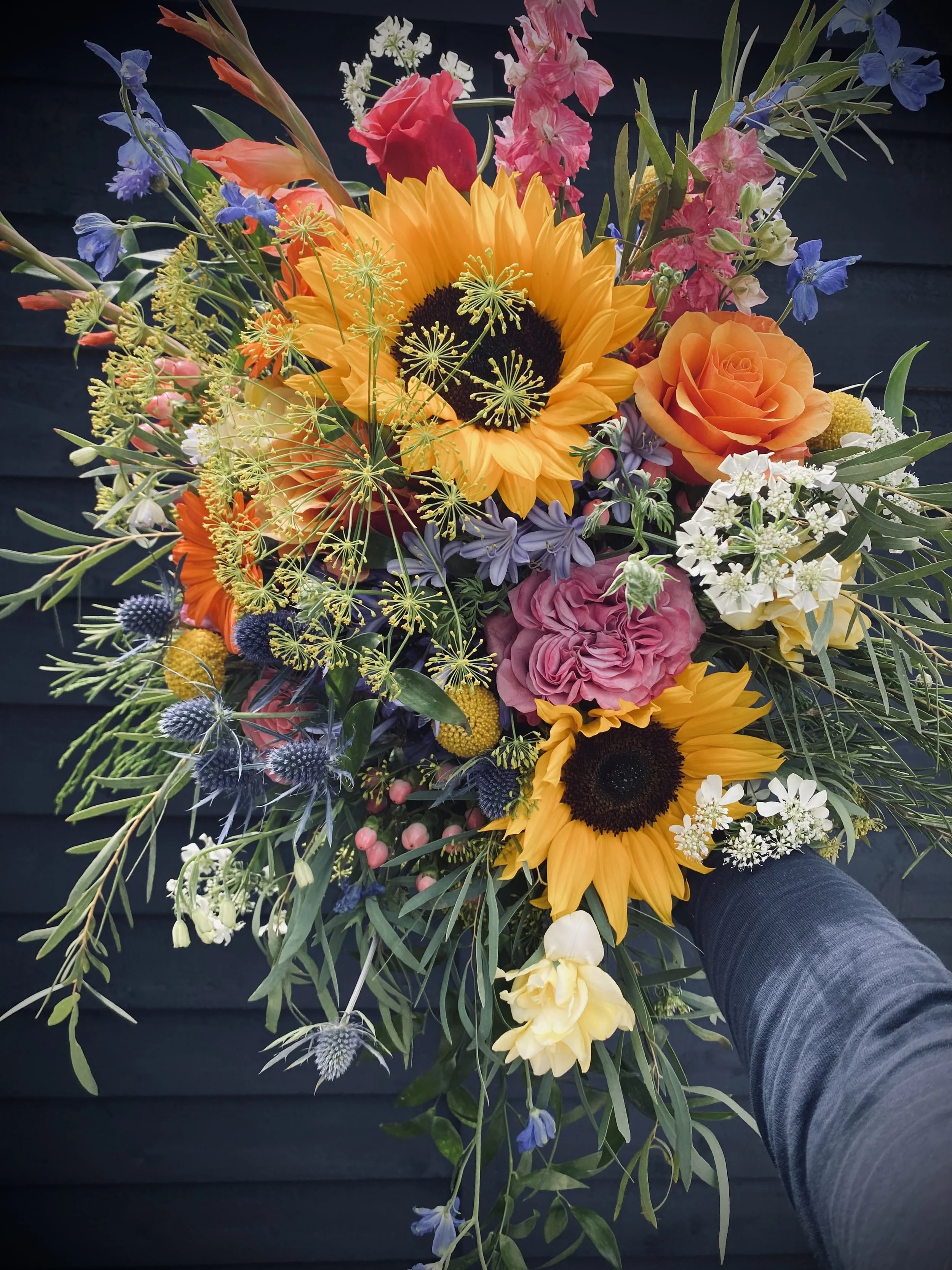 A vibrant bouquet with sunflowers, roses, and various colorful wildflowers held by a person wearing a dark sleeve.