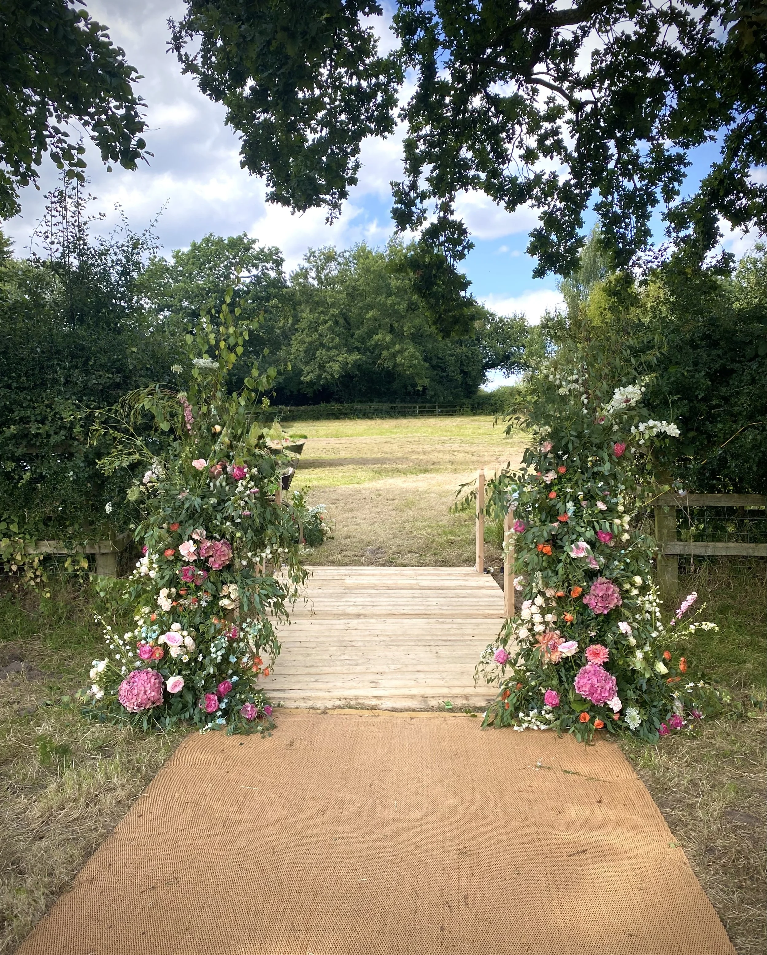 A decorated wooden wedding arch with pink, white, and red flowers at the entrance of a grassy outdoor venue, surrounded by trees.