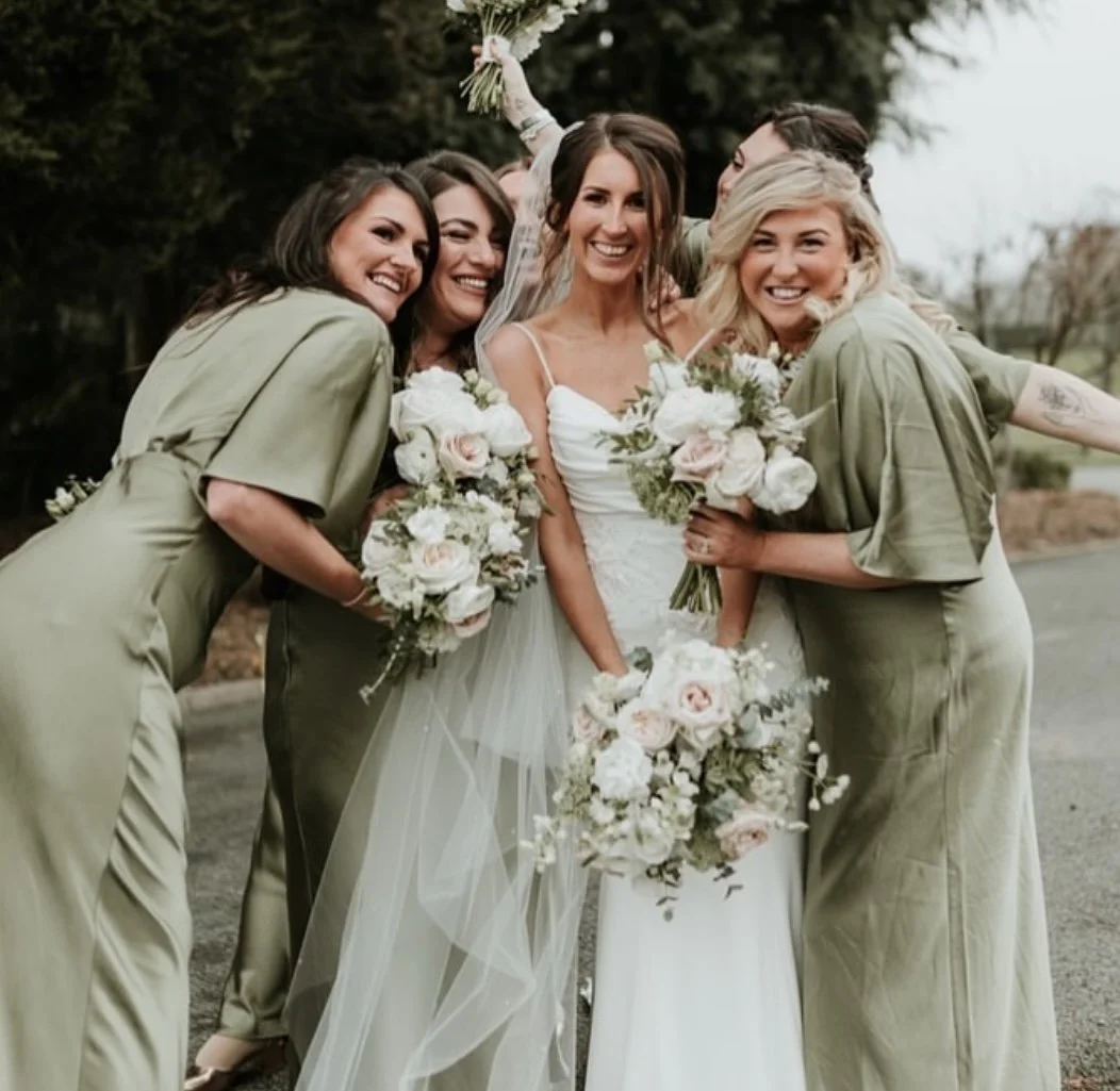 A group of women, including a bride in a white wedding dress holding a bouquet, celebrating outdoors with friends.