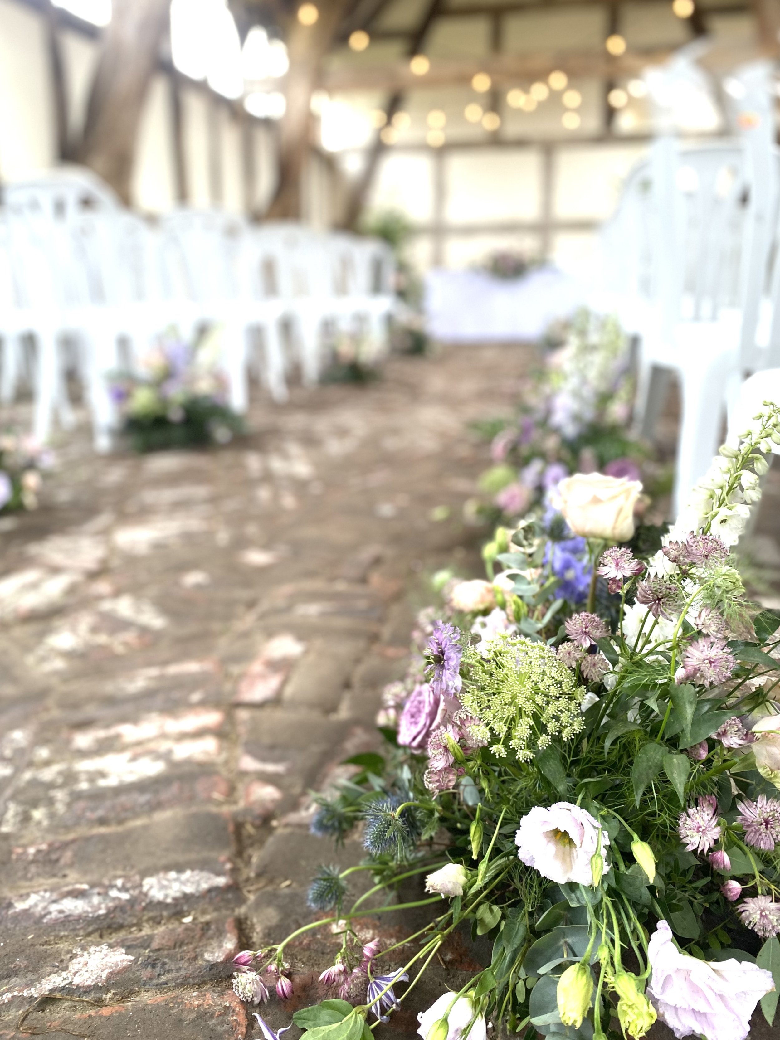 A wedding aisle decorated with pastel-colored flowers along a cobblestone path under string lights, leading to an altar at the back of the event space.
