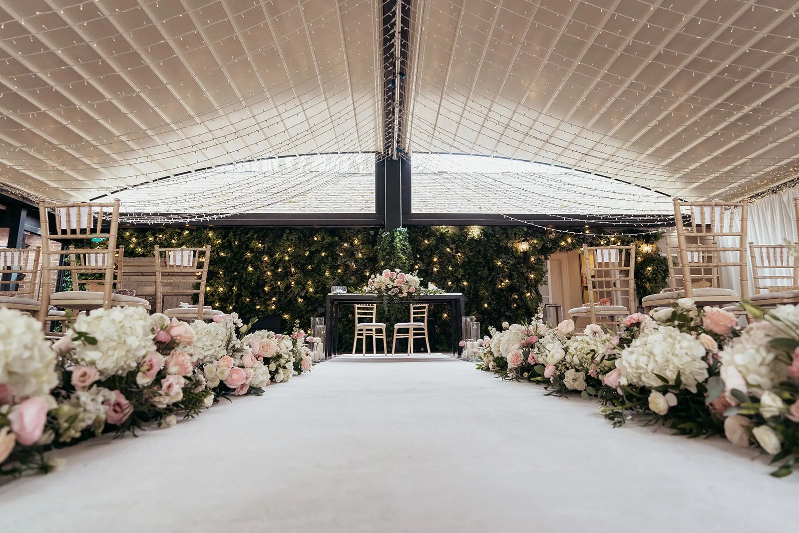 Wedding ceremony setup with floral arrangements along an aisle, chairs on either side, a black table with a floral centerpiece at the front, and a decorated ceiling with string lights.