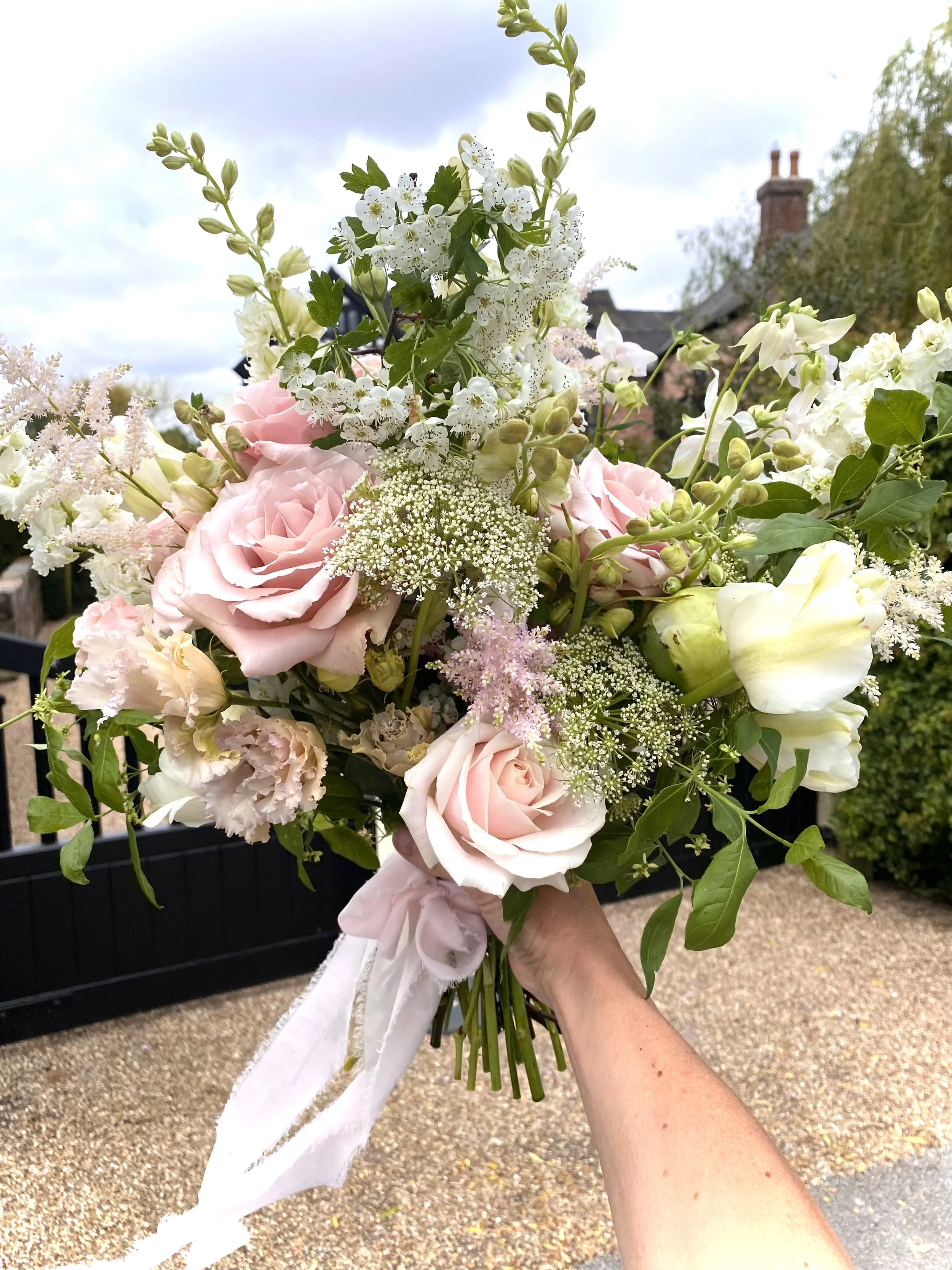 A person holding a bouquet of mixed pink, white, and cream flowers outside, with a house and cloudy sky in the background.
