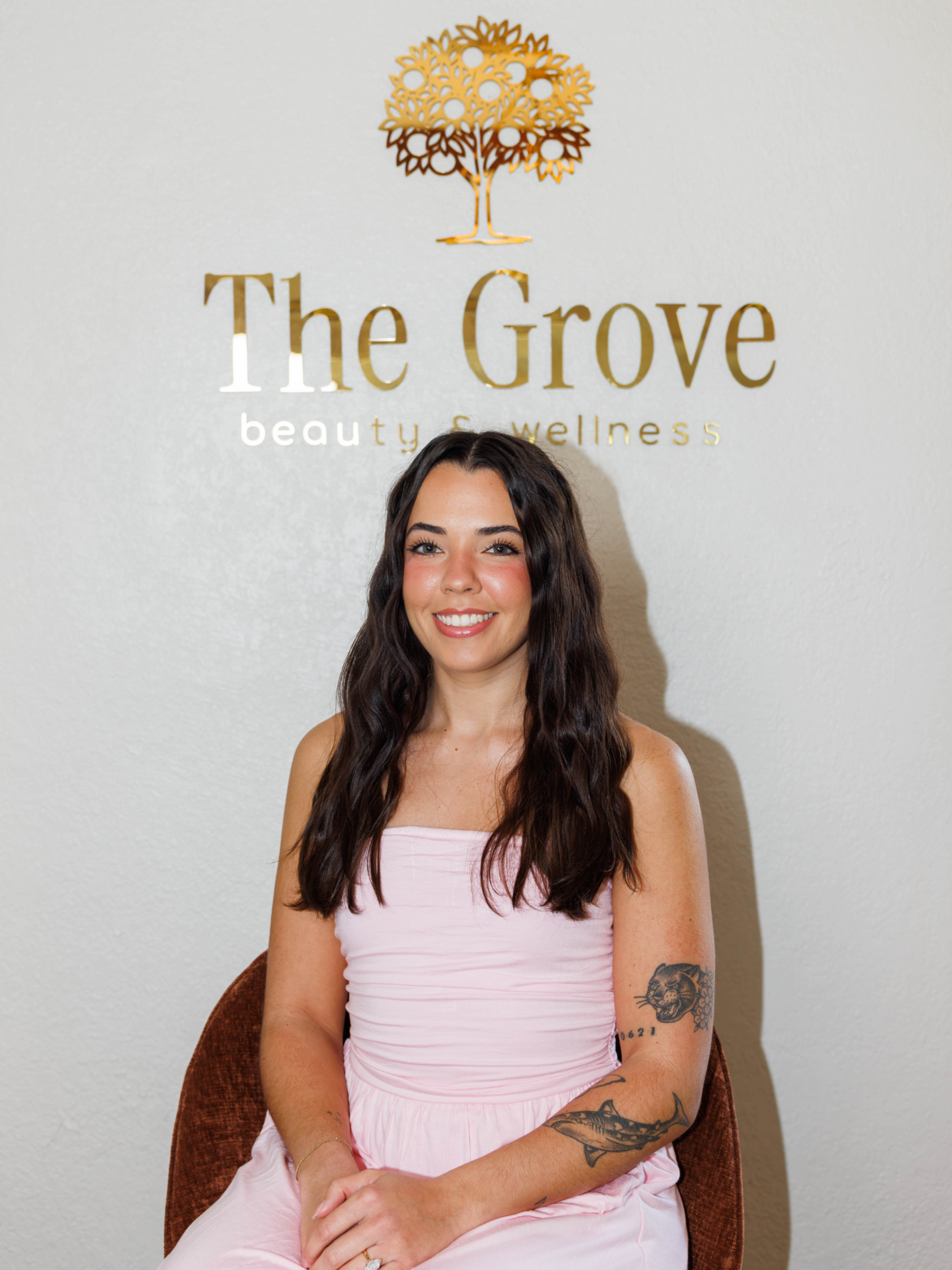 A smiling woman with long dark hair and tattoos, wearing a pink dress, sitting in front of a wall with a sign that reads 'The Grove beauty & wellness' and features a gold tree logo.