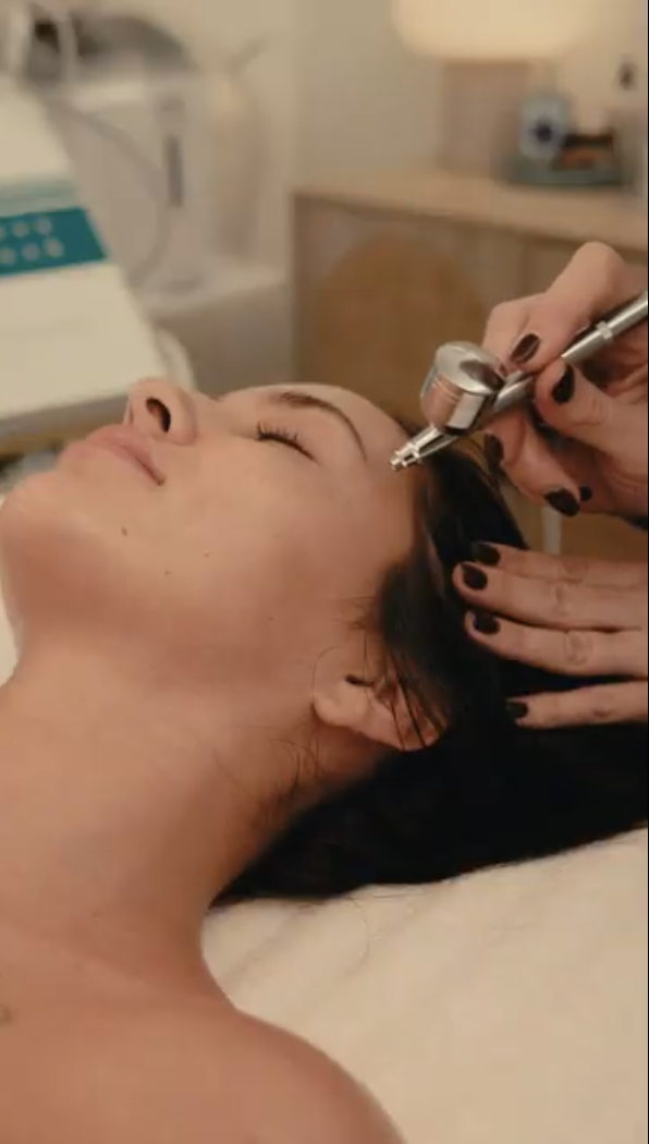 A woman is lying on a bed with eyes closed while receiving a cosmetic injection in her forehead from a professional. The setting appears to be a clinic or spa.