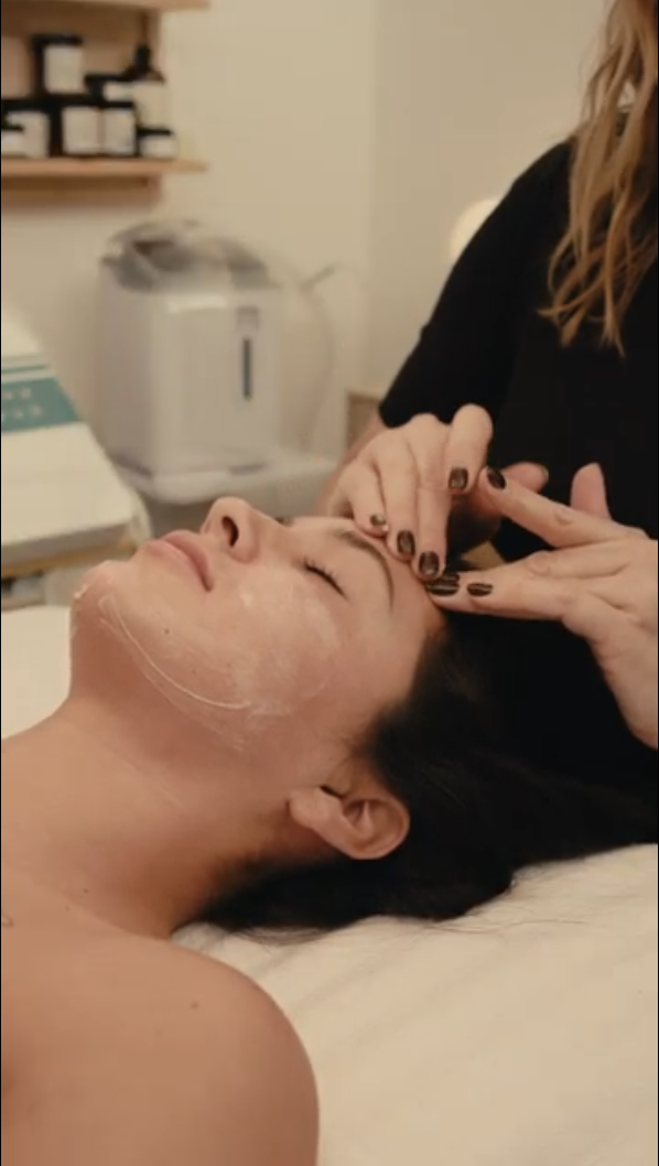 A woman receives a facial treatment from a professional in a spa setting, lying down with a relaxing expression on her face.