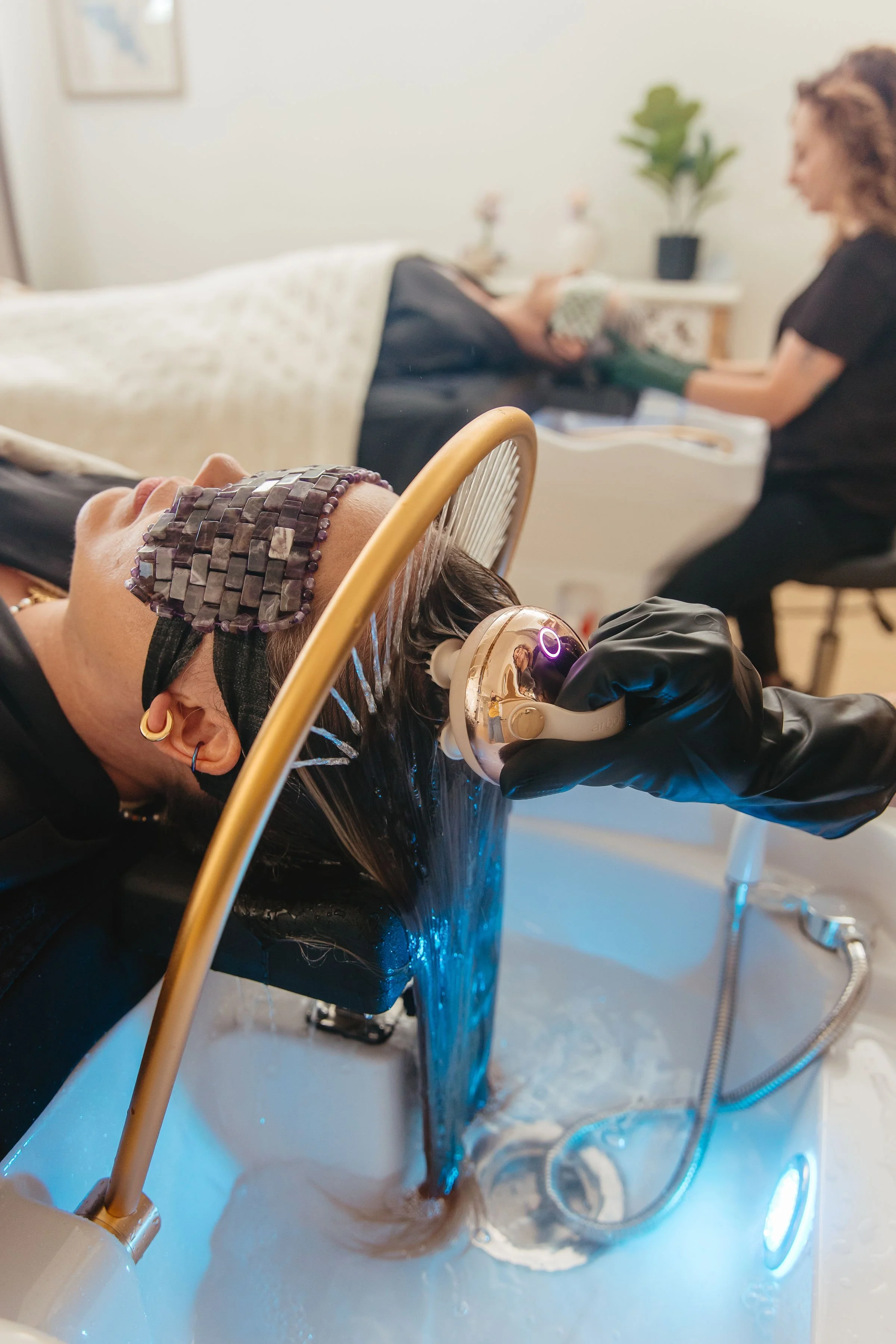 A woman with a headband reclining in a hair treatment basin, receiving a scalp treatment from a professional wearing gloves, as water flows over her hair.