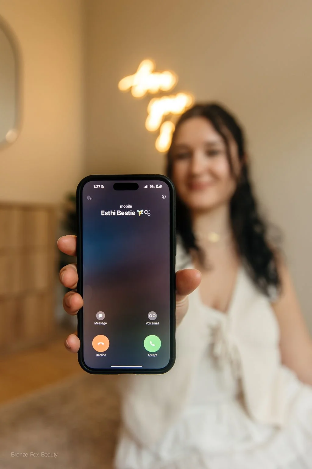 A woman with dark hair and light skin holding a smartphone with a live call from someone named Esthi Bestie, smiling in the background.