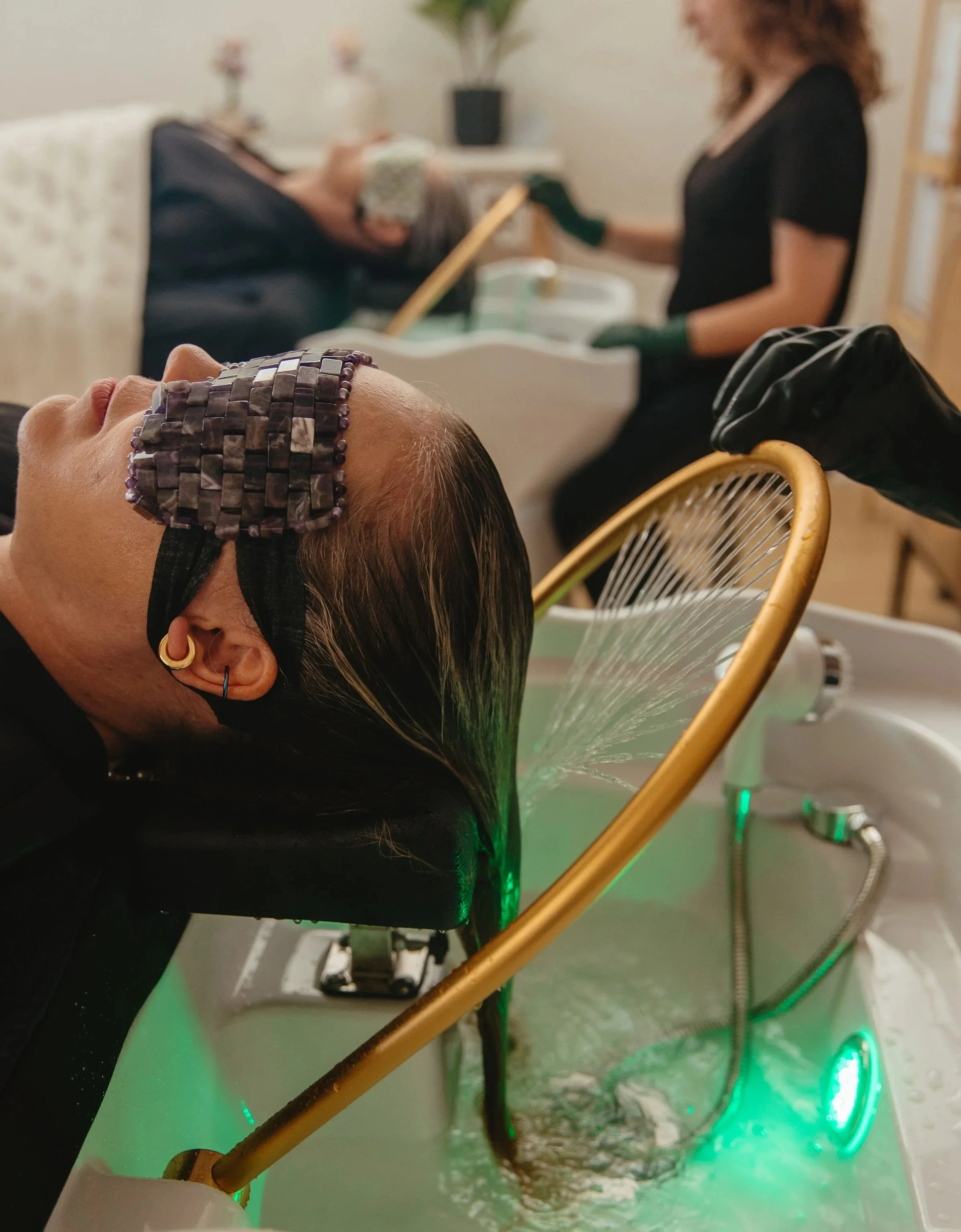 A woman with colorful hair and jewelry gets her hair washed at a salon sink, with her head tilted back in a black salon chair and a stylist in the background rinsing her hair.